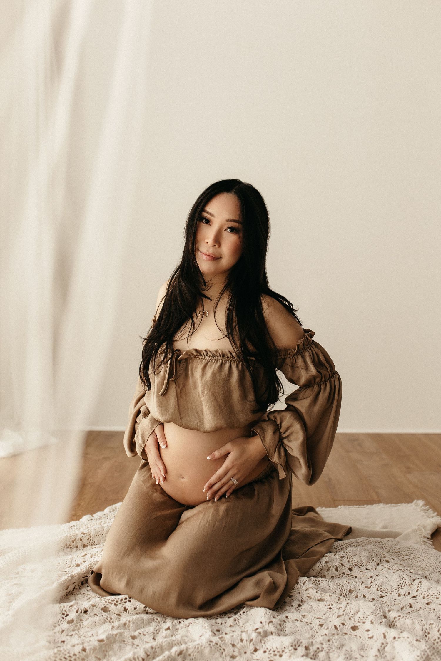 Pregnant woman in beige off-shoulder dress kneeling on floor, cradling baby bump in a serene studio setting.