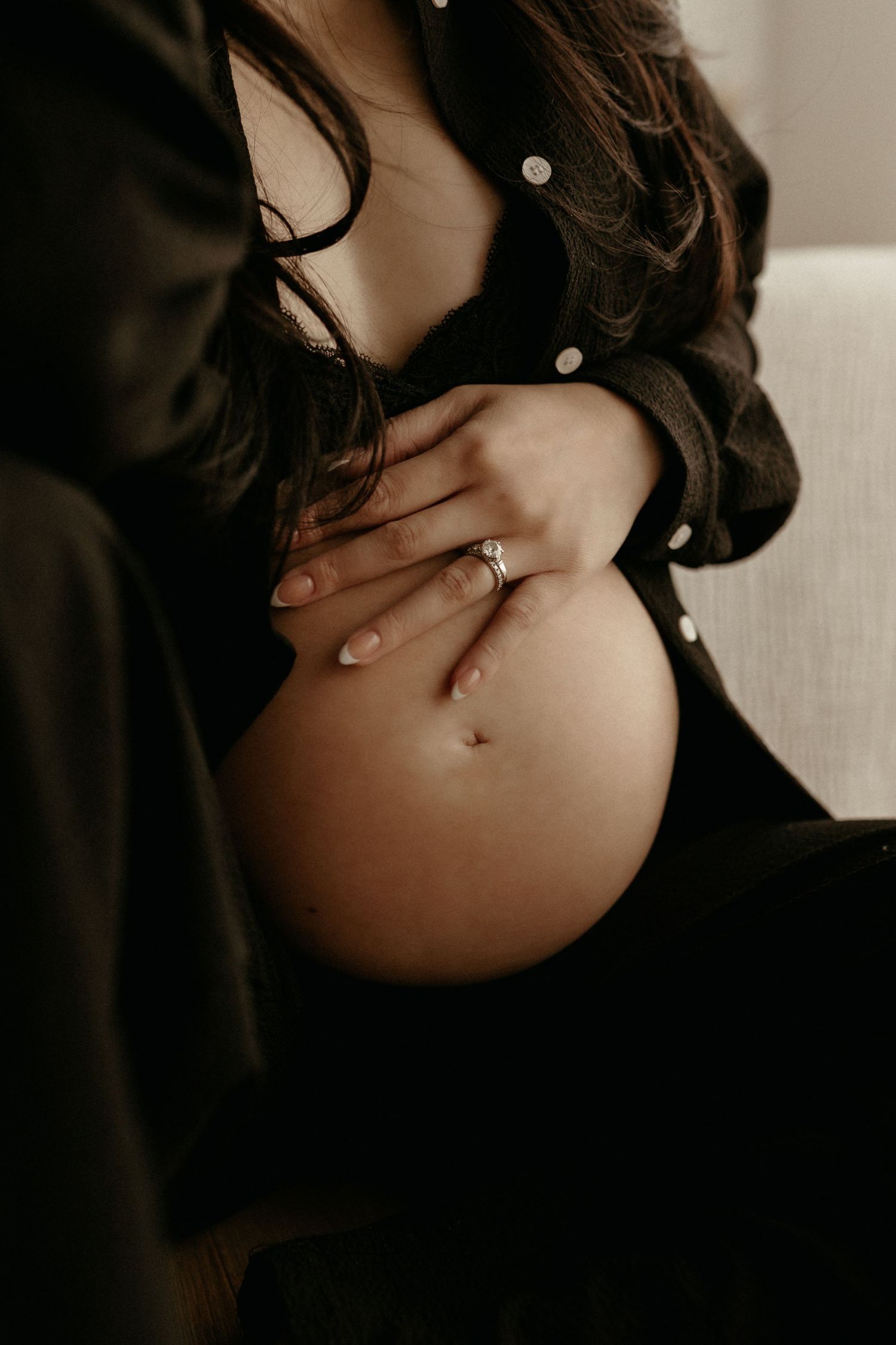 Pregnant woman in black shirt cradling bare baby bump, wearing engagement ring, soft moody lighting.