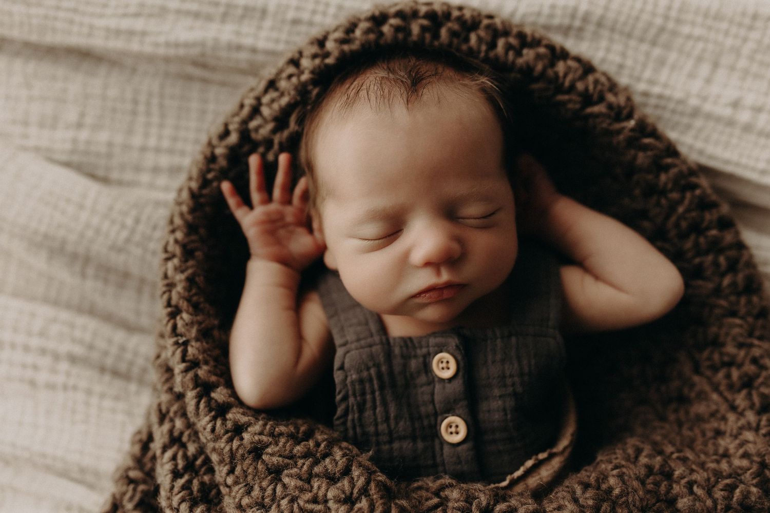 Peaceful newborn baby sleeping curled in a brown knit bowl, wearing a linen romper, perfect newborn photography pose.