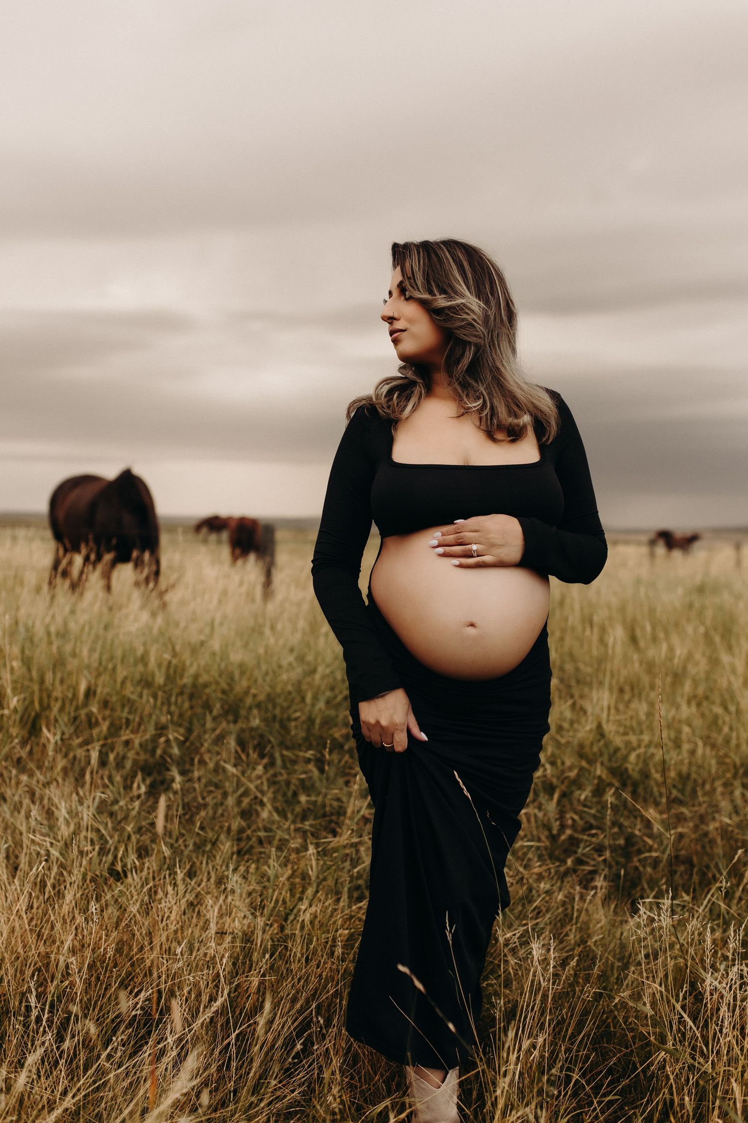 Pregnant woman in black dress cradling baby bump in grassy field with horses grazing under cloudy sky.