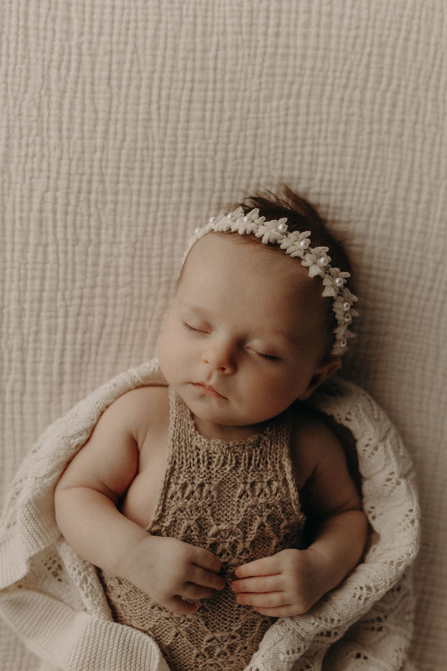 Sleeping newborn baby girl wearing a delicate floral headband and knit romper, wrapped in a soft cream blanket.