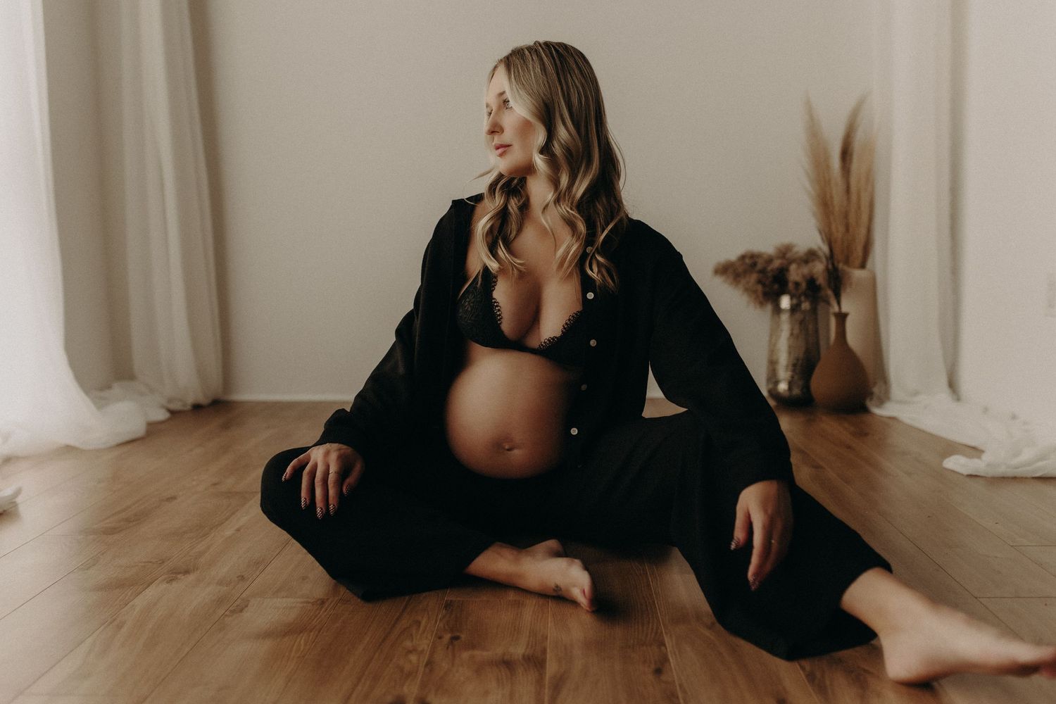 Pregnant woman in black outfit sitting gracefully on wooden floor during maternity photoshoot in bright studio.