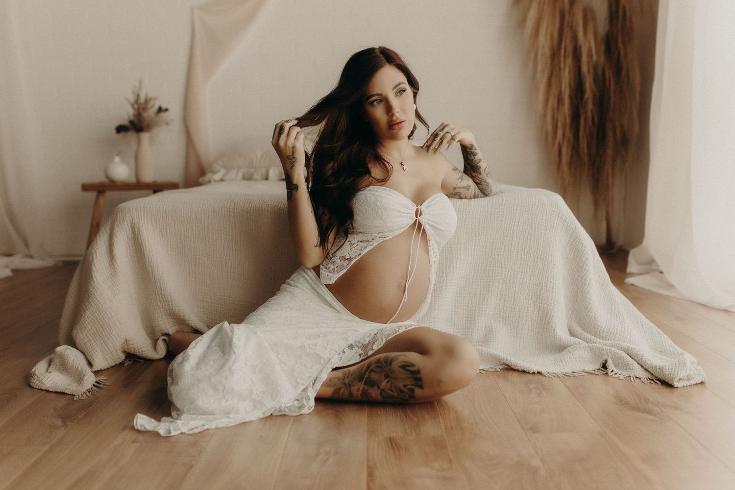 Pregnant woman in white lace outfit sitting gracefully on hardwood floor during elegant maternity photo session.