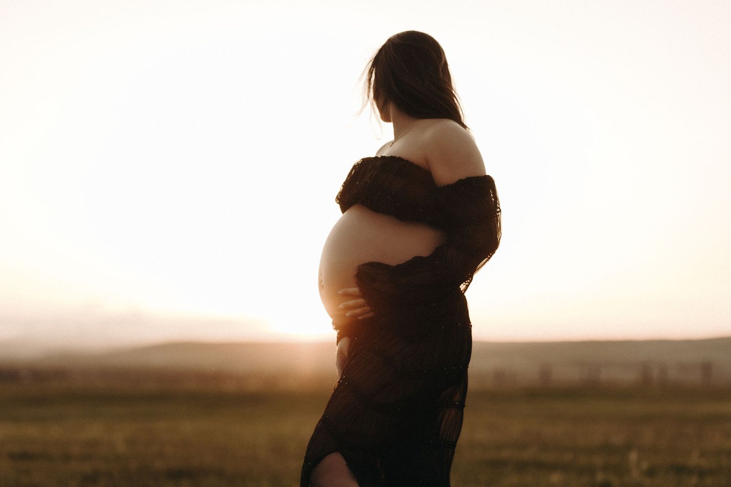 Pregnant woman cradling her bare baby bump outdoors at golden sunset in a flowing black dress.