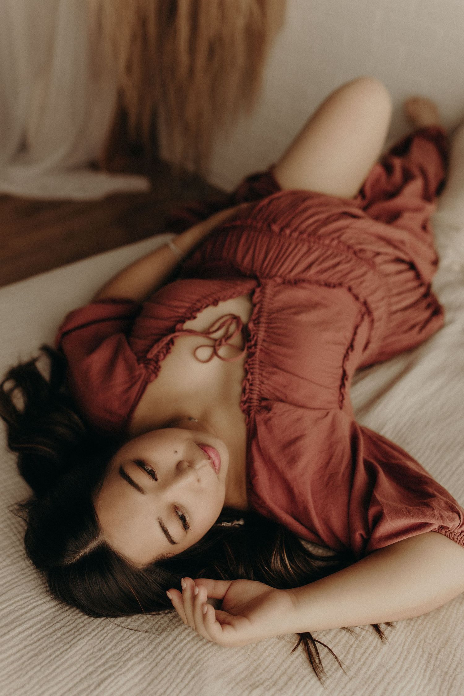 Woman in rust-colored dress lying on bed with eyes closed, dark hair spread out, in soft natural light.