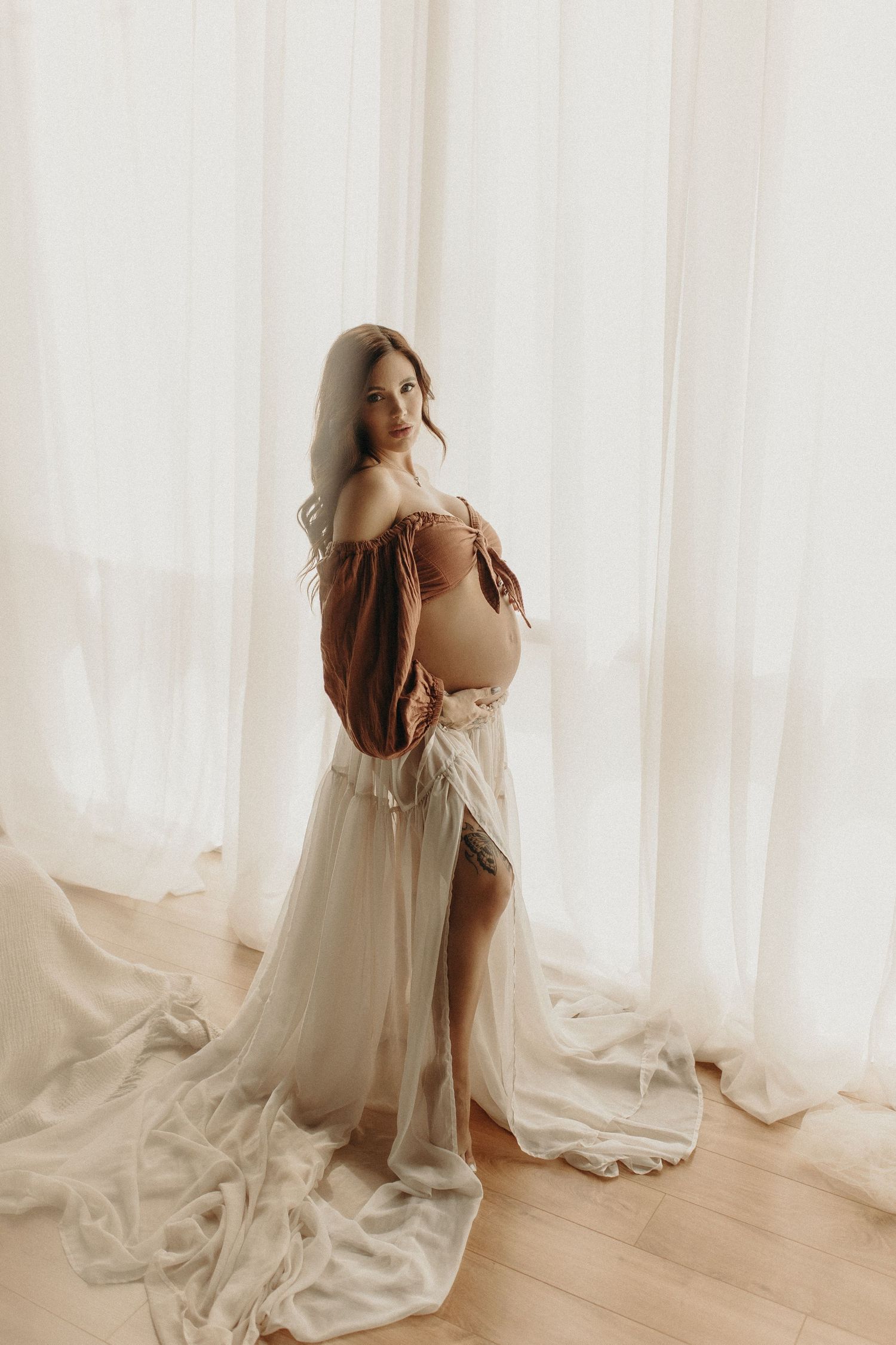 Pregnant woman in rust top and flowing white tulle skirt stands gracefully by sheer curtains in soft natural light.