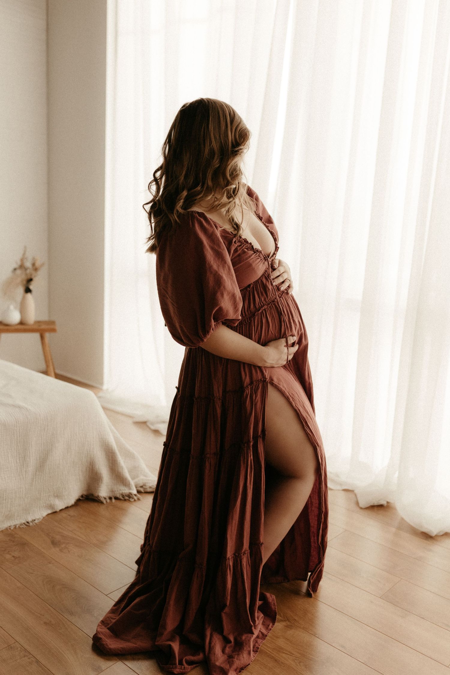 Pregnant woman in flowing rust-colored maternity gown cradling baby bump by sheer curtained window.