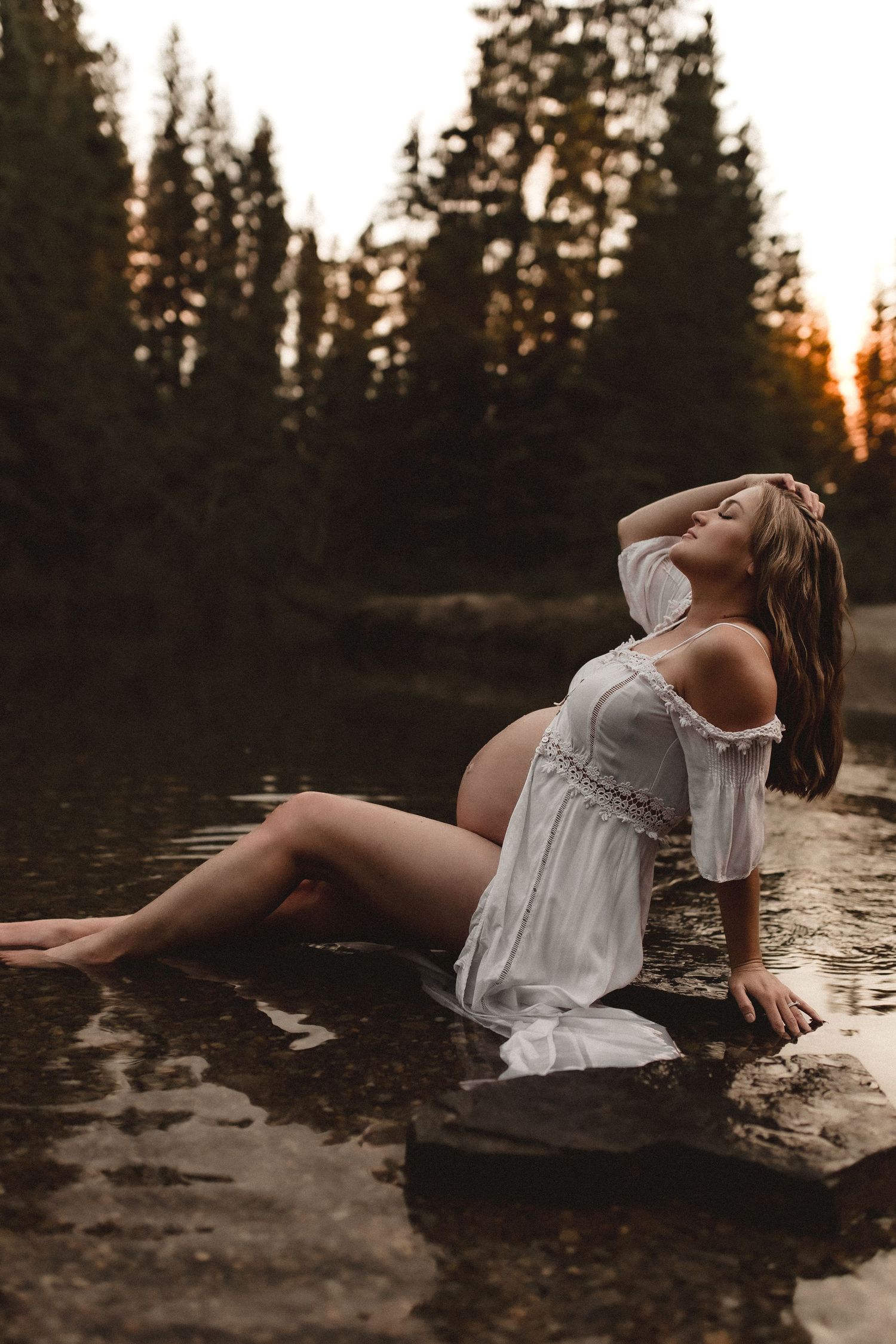 Pregnant woman in white dress sitting on rocks by a forest lake at golden hour sunset.
