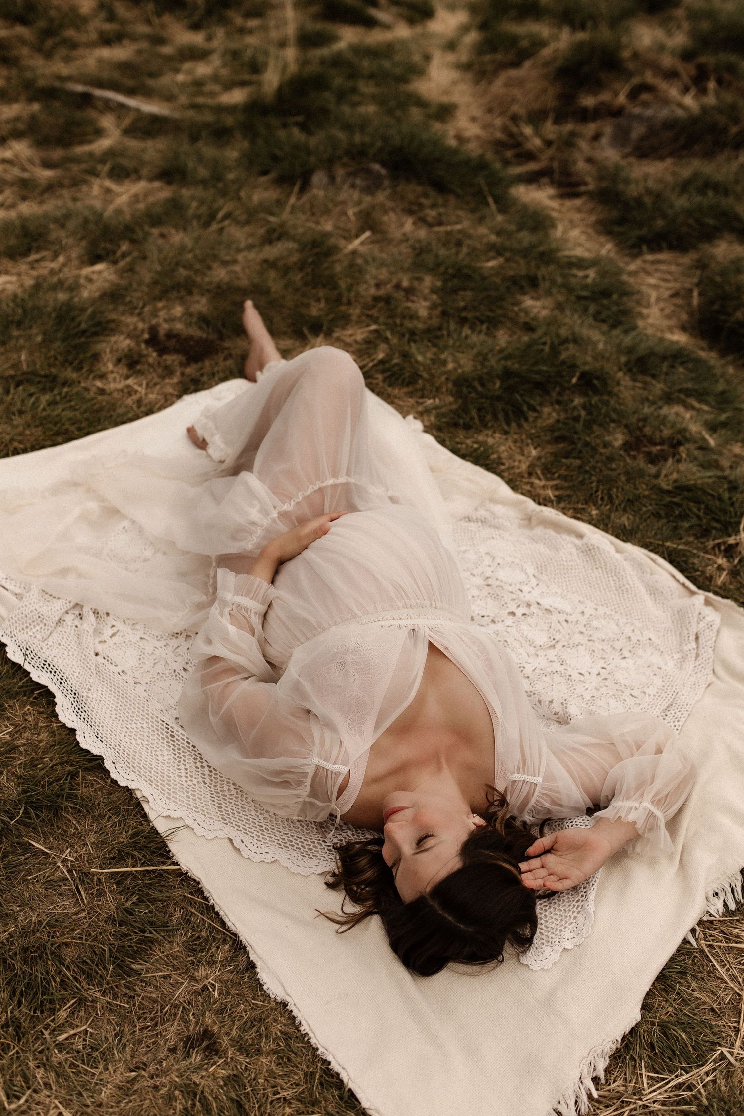 Pregnant woman in sheer white lace robe lying on cream blanket on grass field.