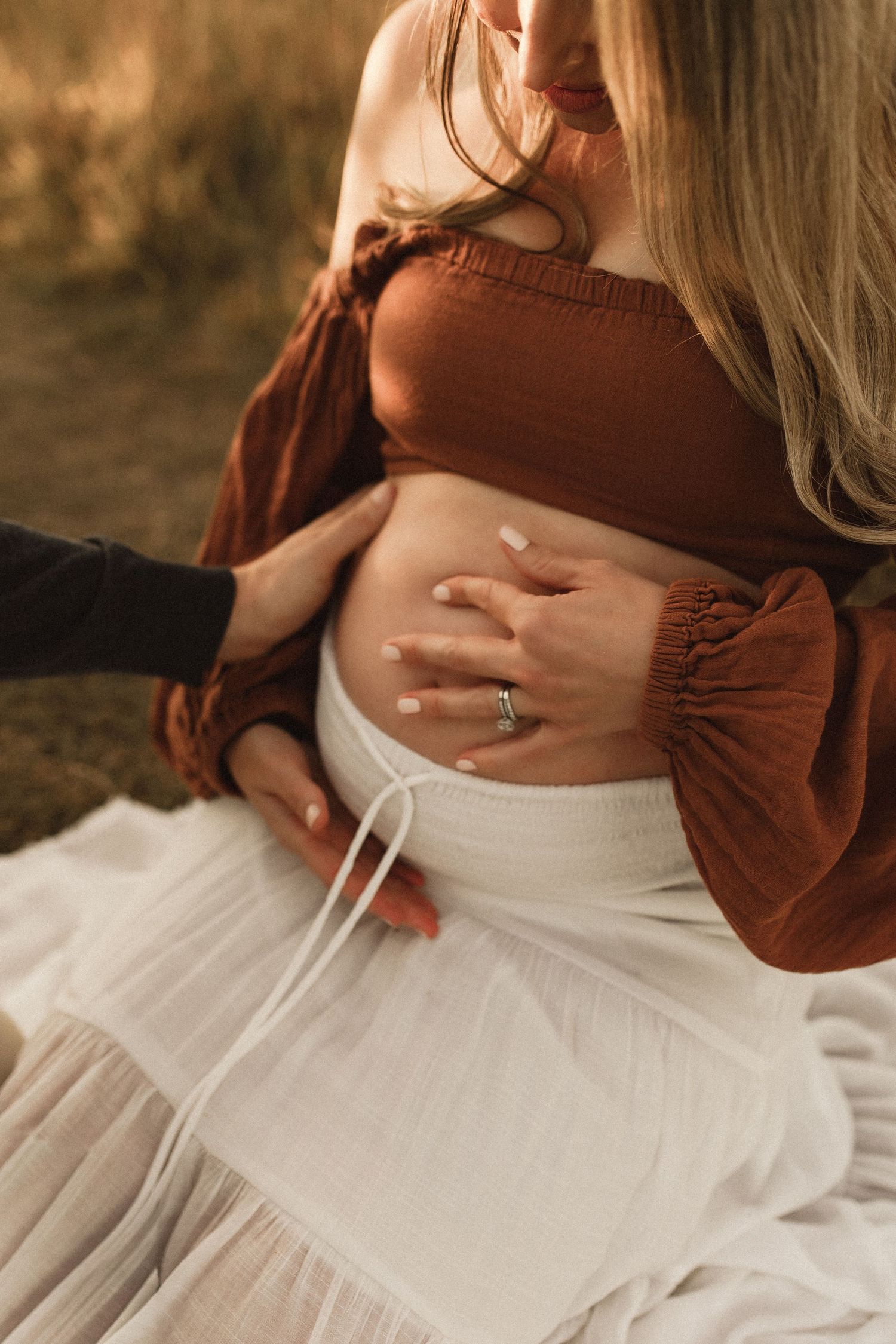 Pregnant woman in rust crop top and white skirt cradling baby bump with partner's hand, outdoor maternity photo.