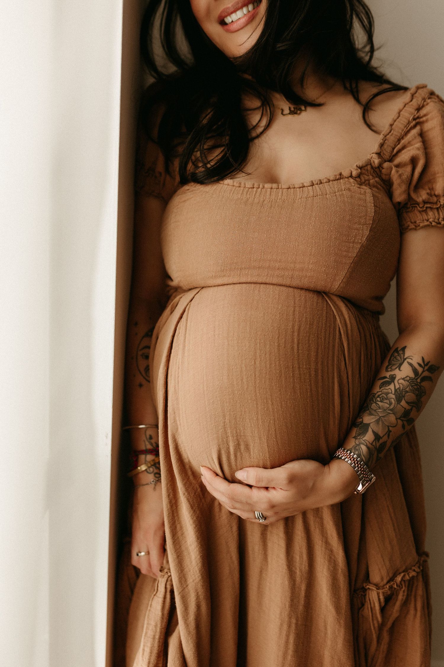 Smiling pregnant woman in tan linen dress cradling baby bump, tattooed arms adorned with silver rings and jewelry.