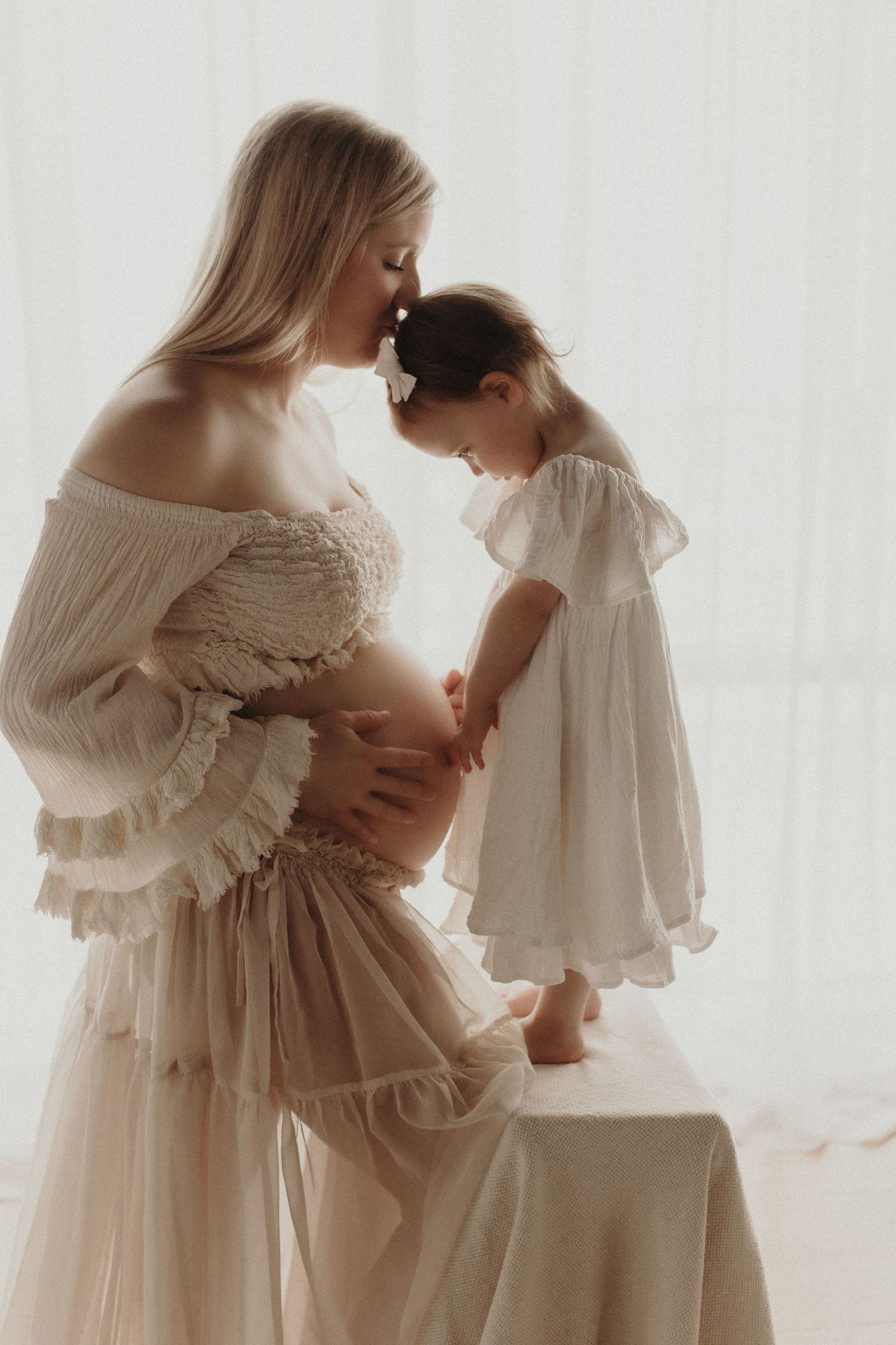 Pregnant woman in beige dress tenderly touching foreheads with toddler girl in white dress near bright window.