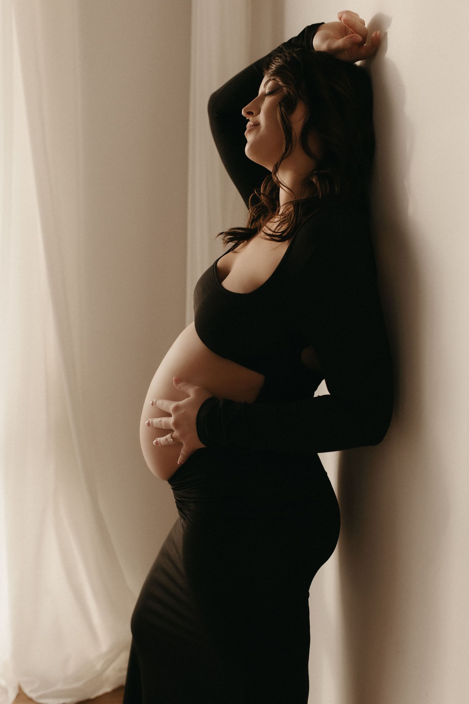 Pregnant woman in black cutout dress cradling baby bump, leaning against wall in soft natural light.