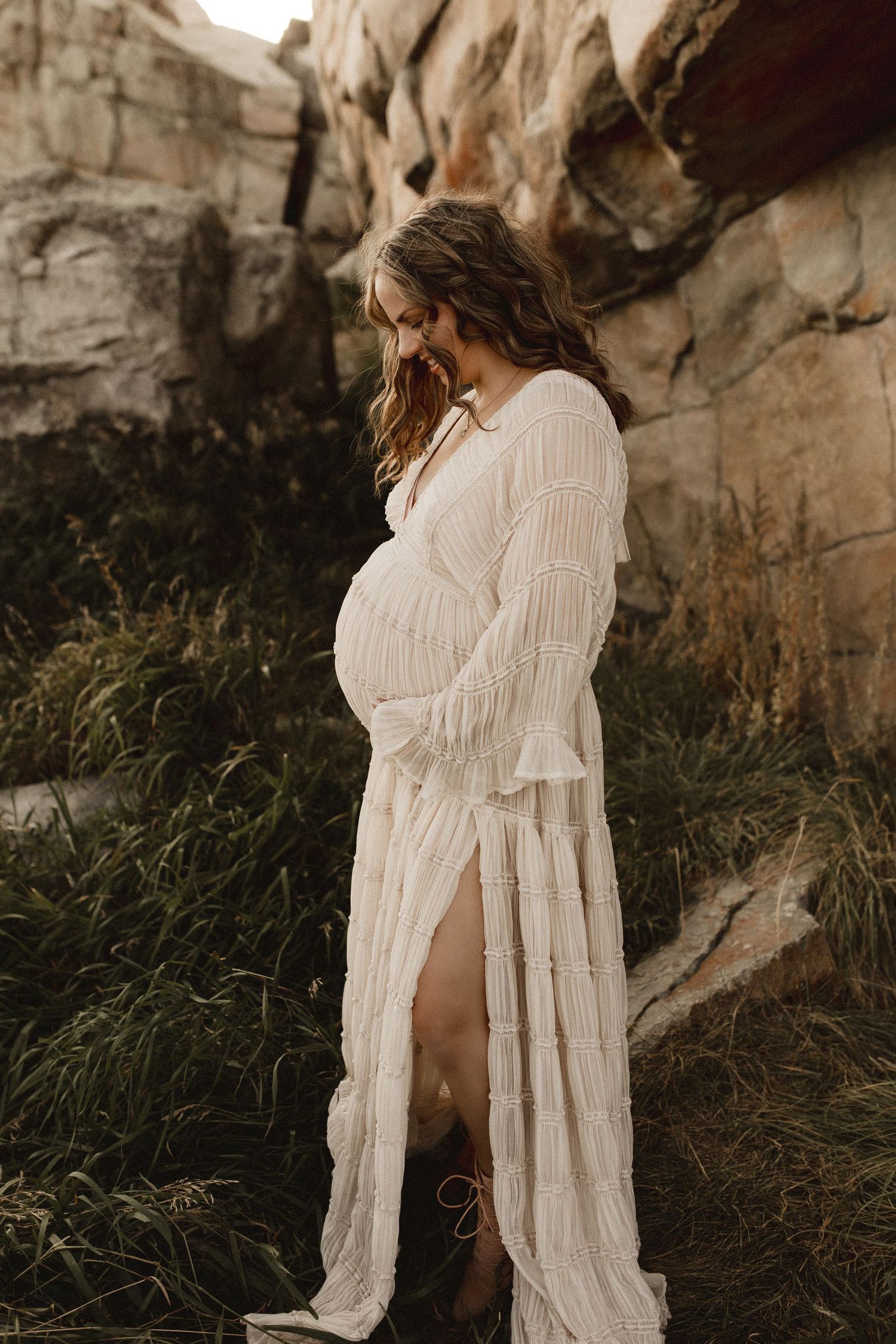 Smiling pregnant woman in white lace maxi dress poses outdoors among rocks and greenery in a boho maternity photo session.