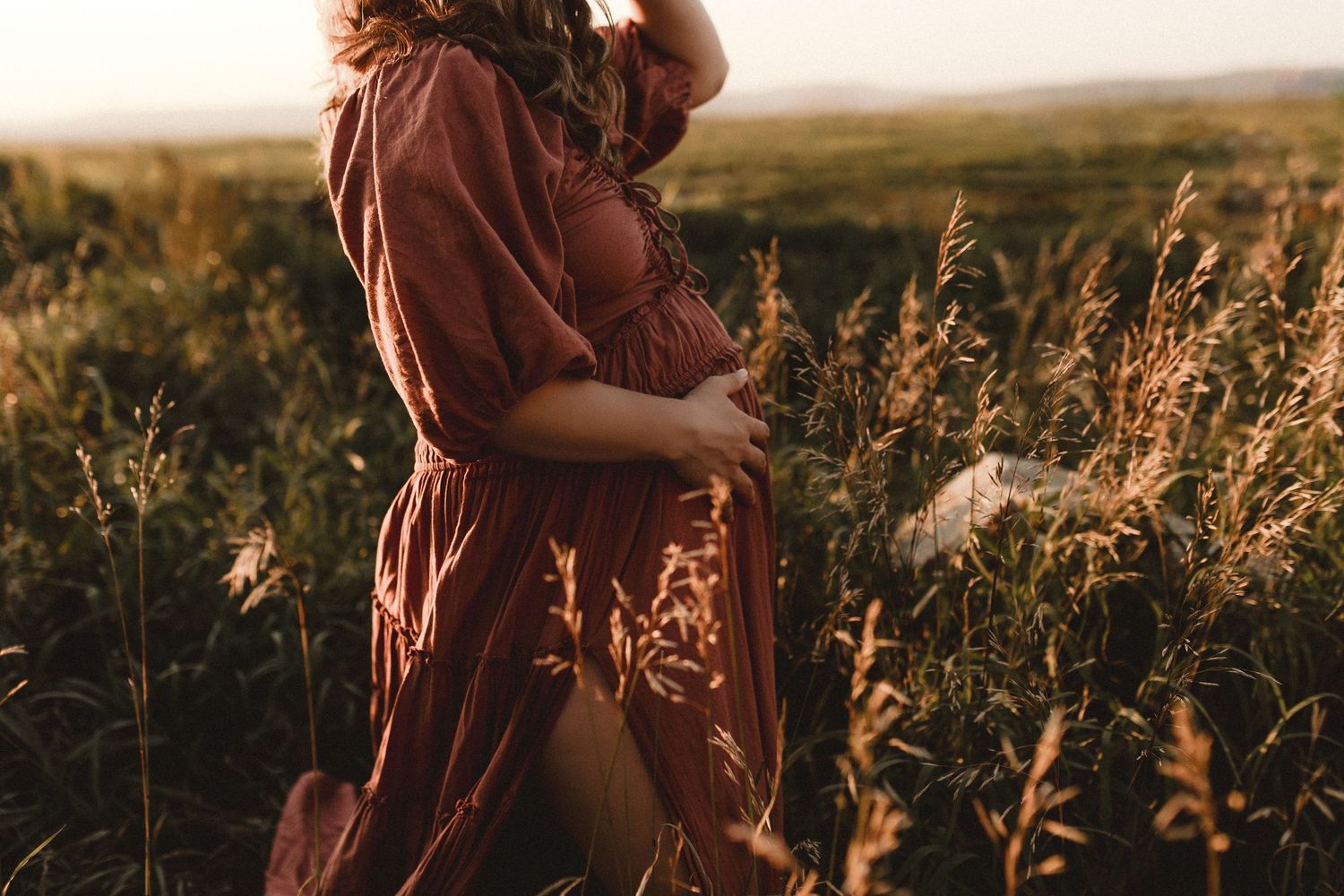 Pregnant woman in rust boho dress cradling baby bump in golden field at sunset.