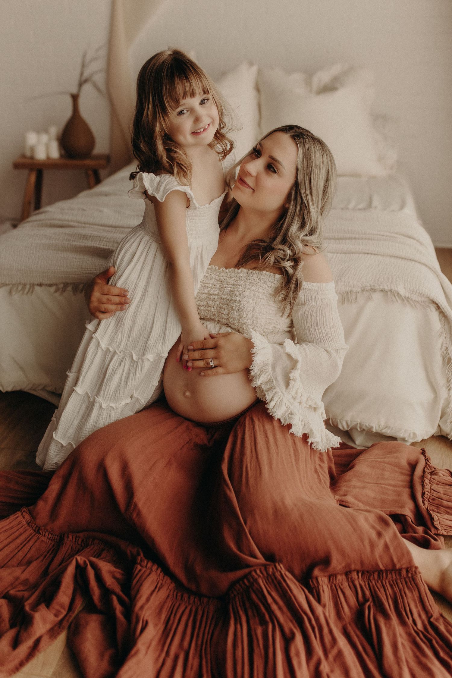 Pregnant woman in white top and rust skirt sits on bed with smiling young daughter in white dress touching her baby bump.