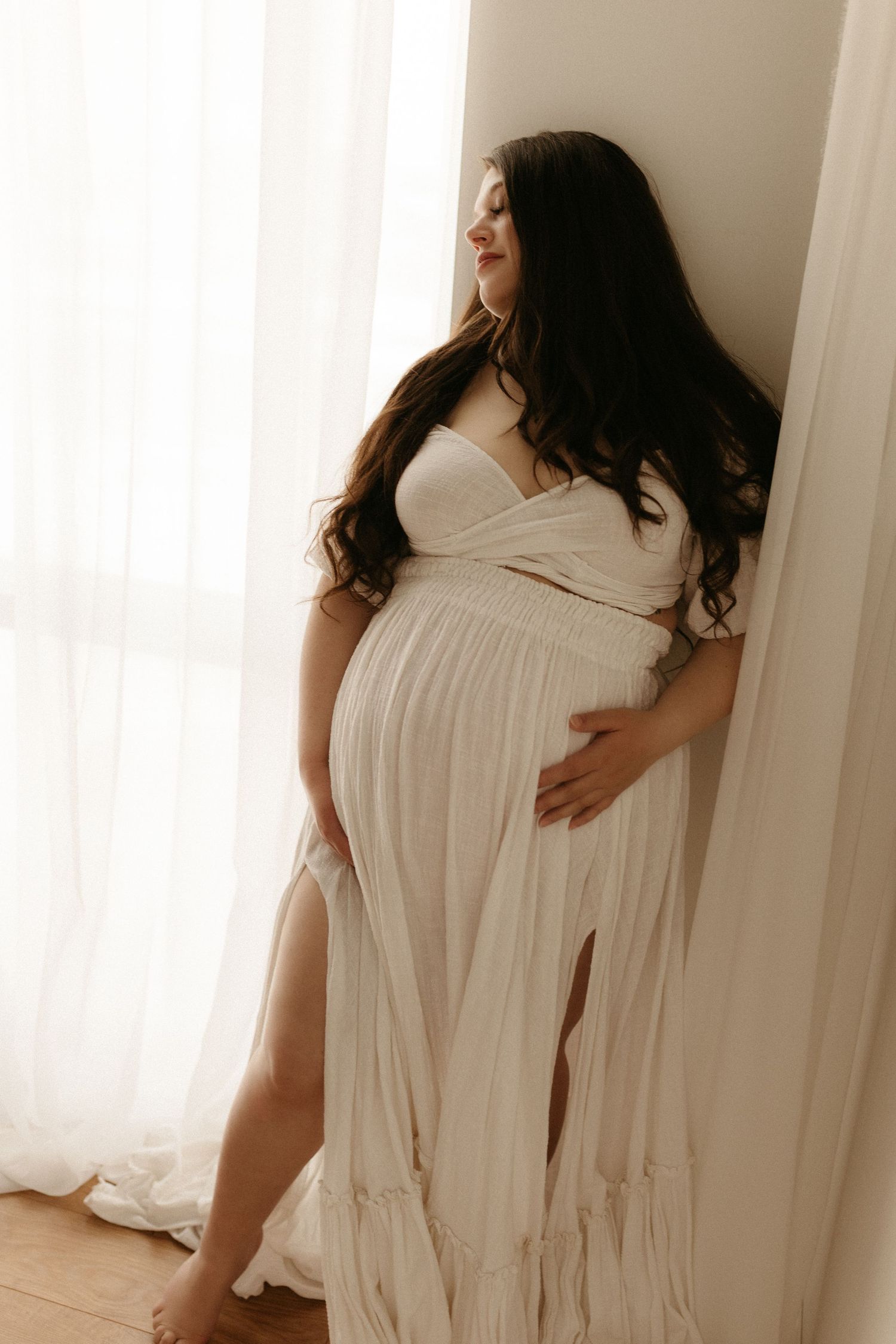 Pregnant woman in white flowing maternity dress leaning against wall by sheer curtained window in soft natural light.