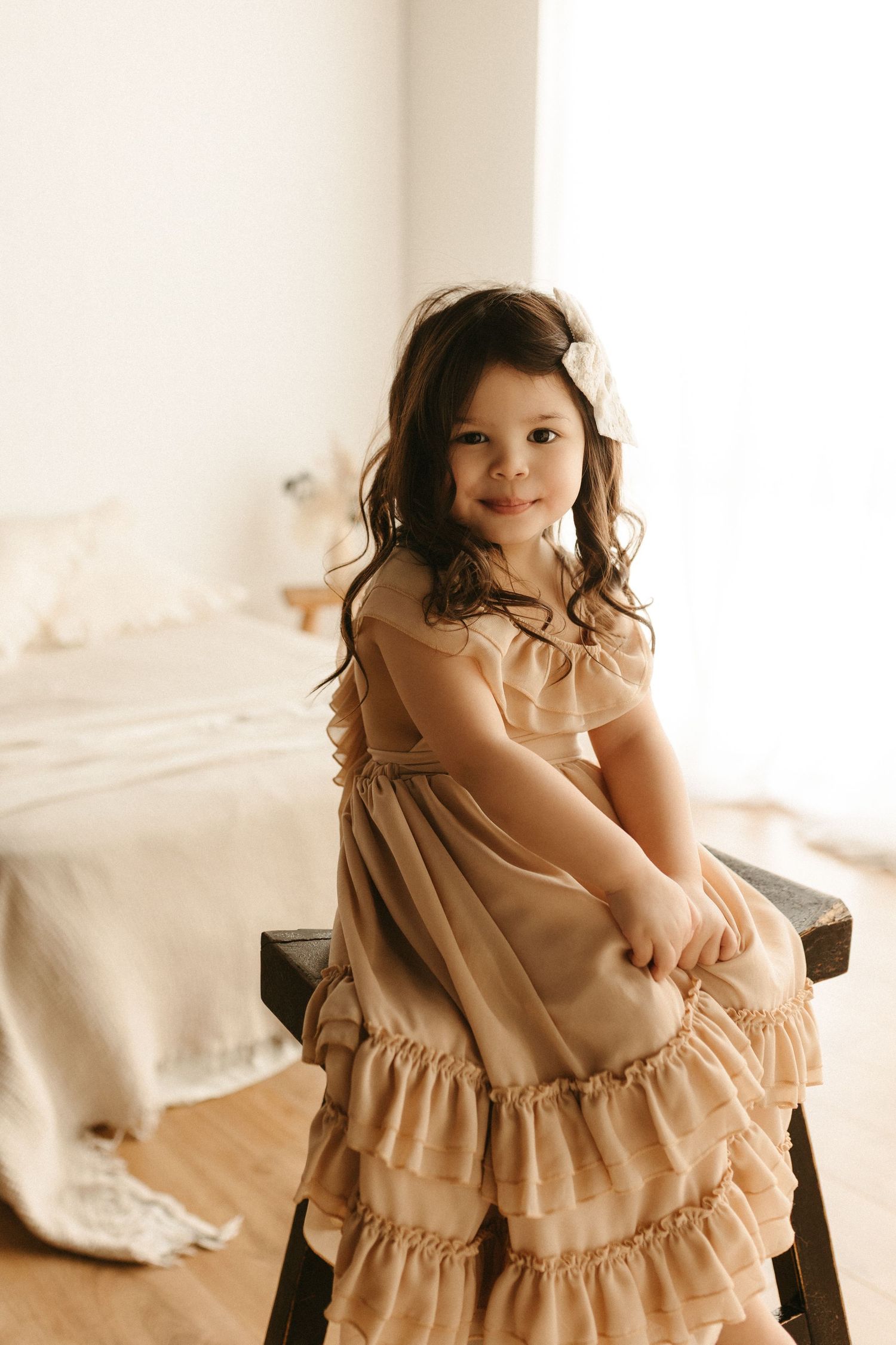 A young girl in a ruffled beige dress with a white bow sits on a bench, smiling in a bright, elegant room.