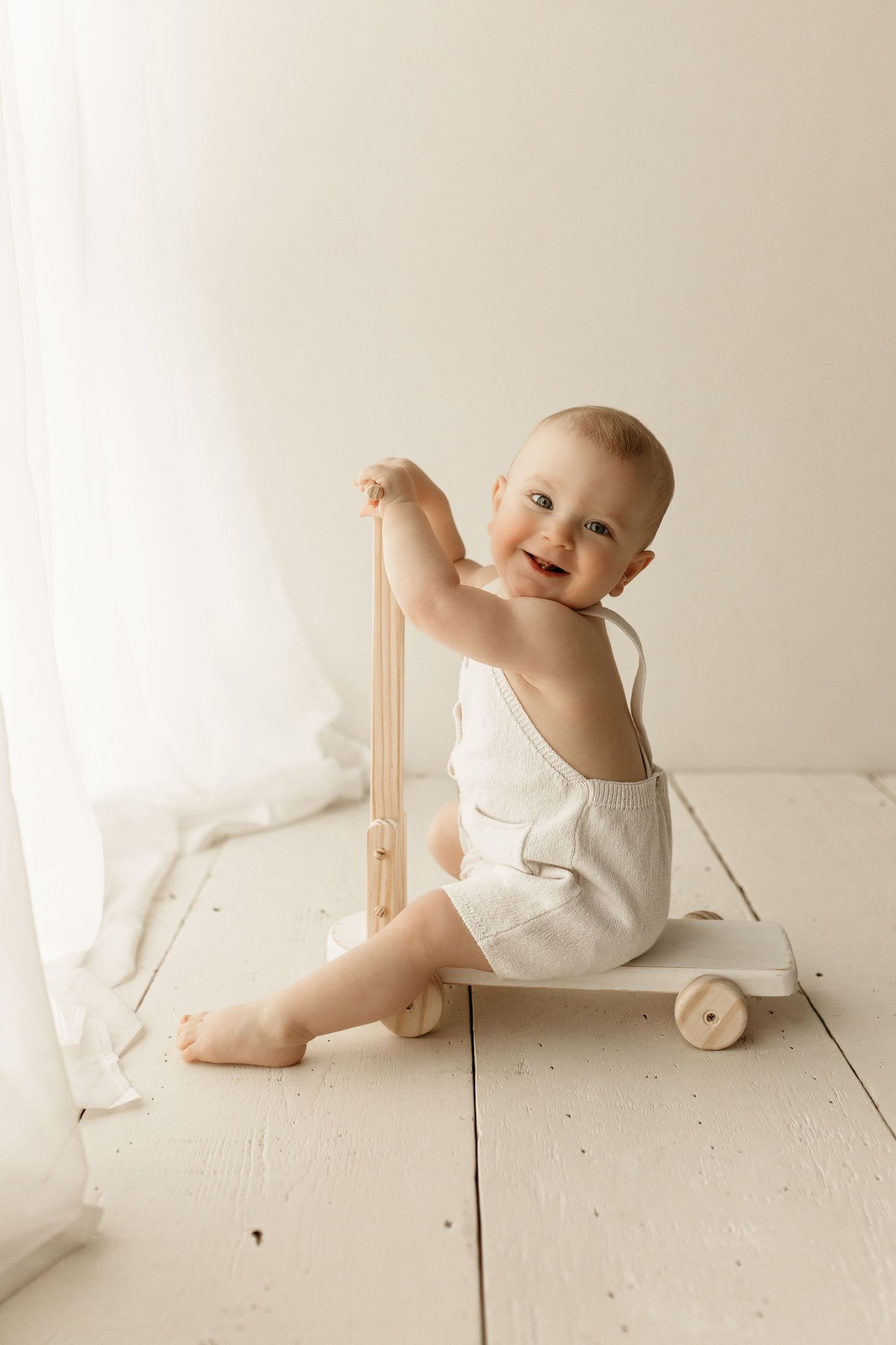 Smiling baby in white romper sitting on wooden floor holding a toy wooden walker cart near sheer curtains.