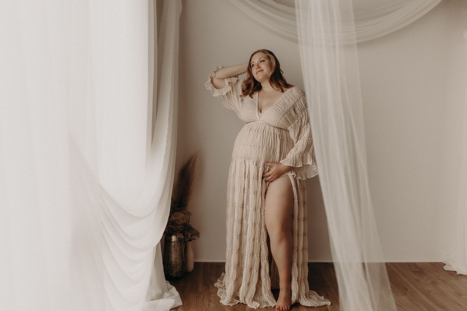 Pregnant woman in elegant lace maternity gown poses gracefully between sheer white curtains in a softly lit studio.
