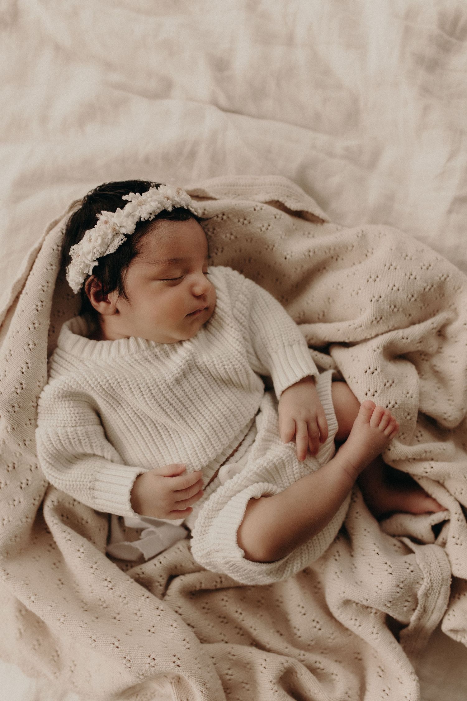 Sleeping newborn baby in cream knit sweater and floral headband, nestled in soft beige blankets.