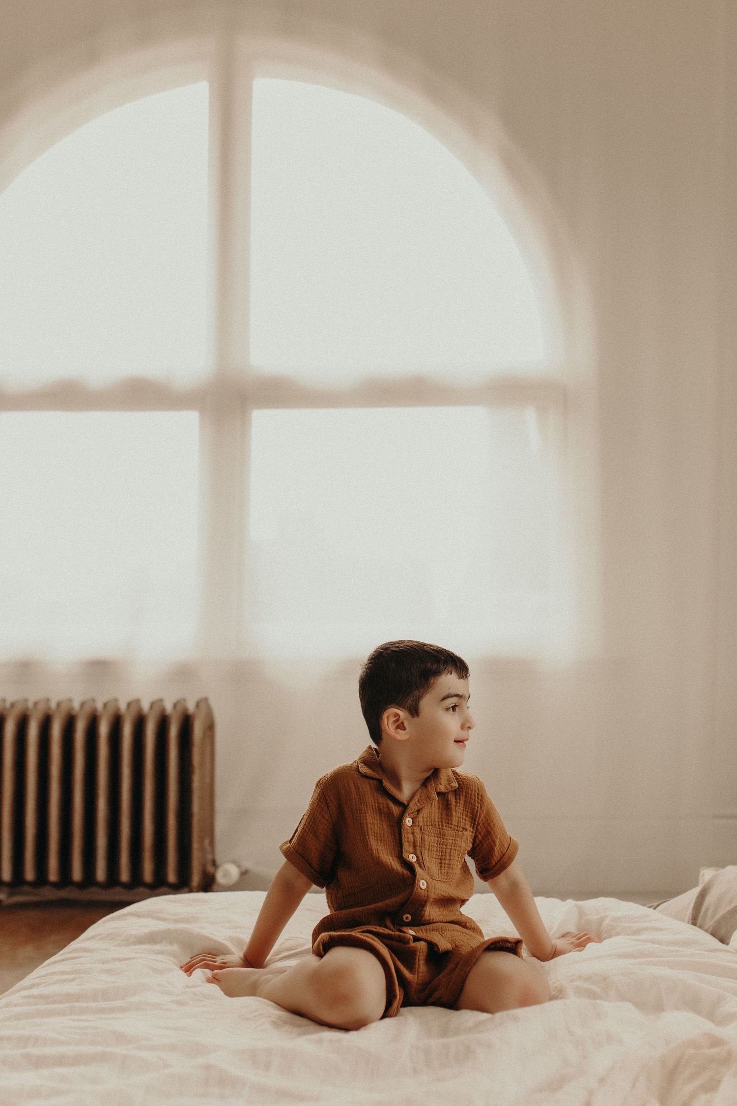 Young boy in brown shorts set sitting on white bed near arched window with radiator in bright room.