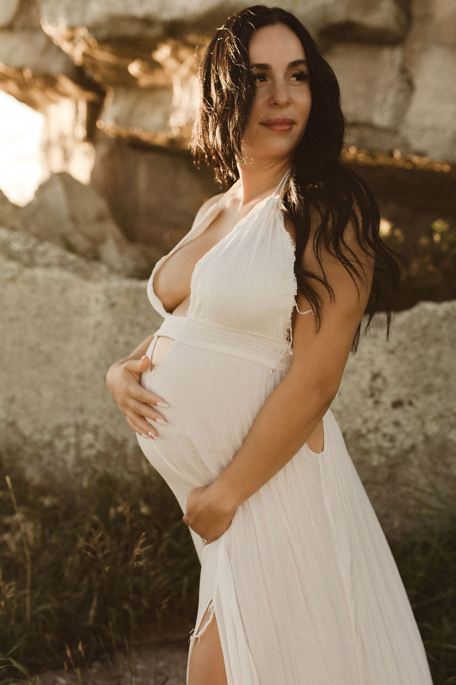 Pregnant woman in white halter dress cradling baby bump outdoors near rocky backdrop in warm golden light.