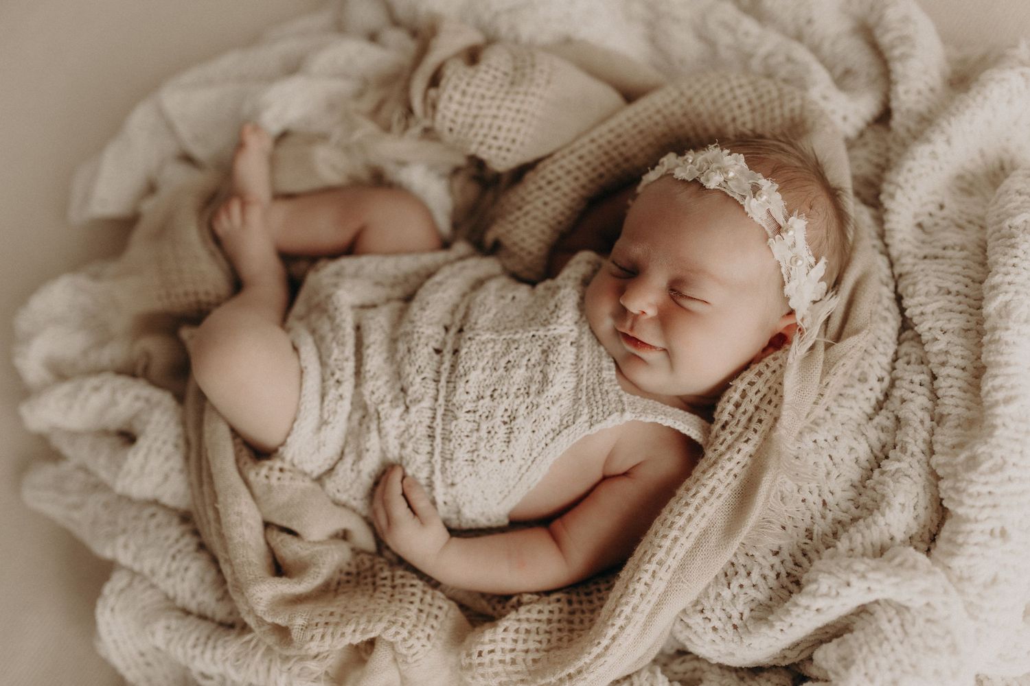 Newborn baby sleeping peacefully in a cream knit blanket nest, wearing a delicate floral headband.