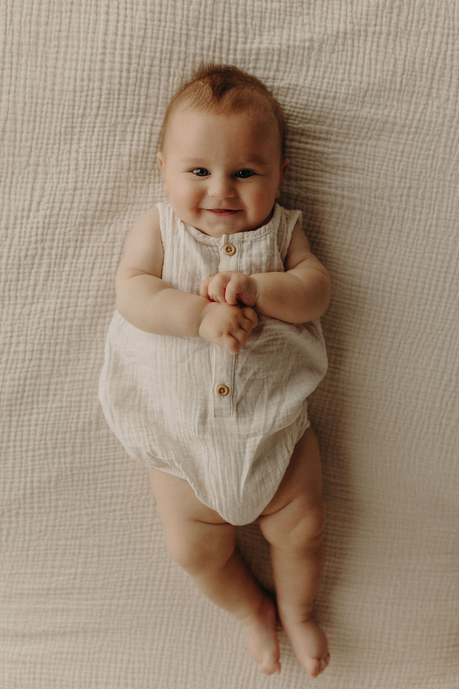 Smiling baby lying on a white textured blanket wearing a beige linen romper with buttons.