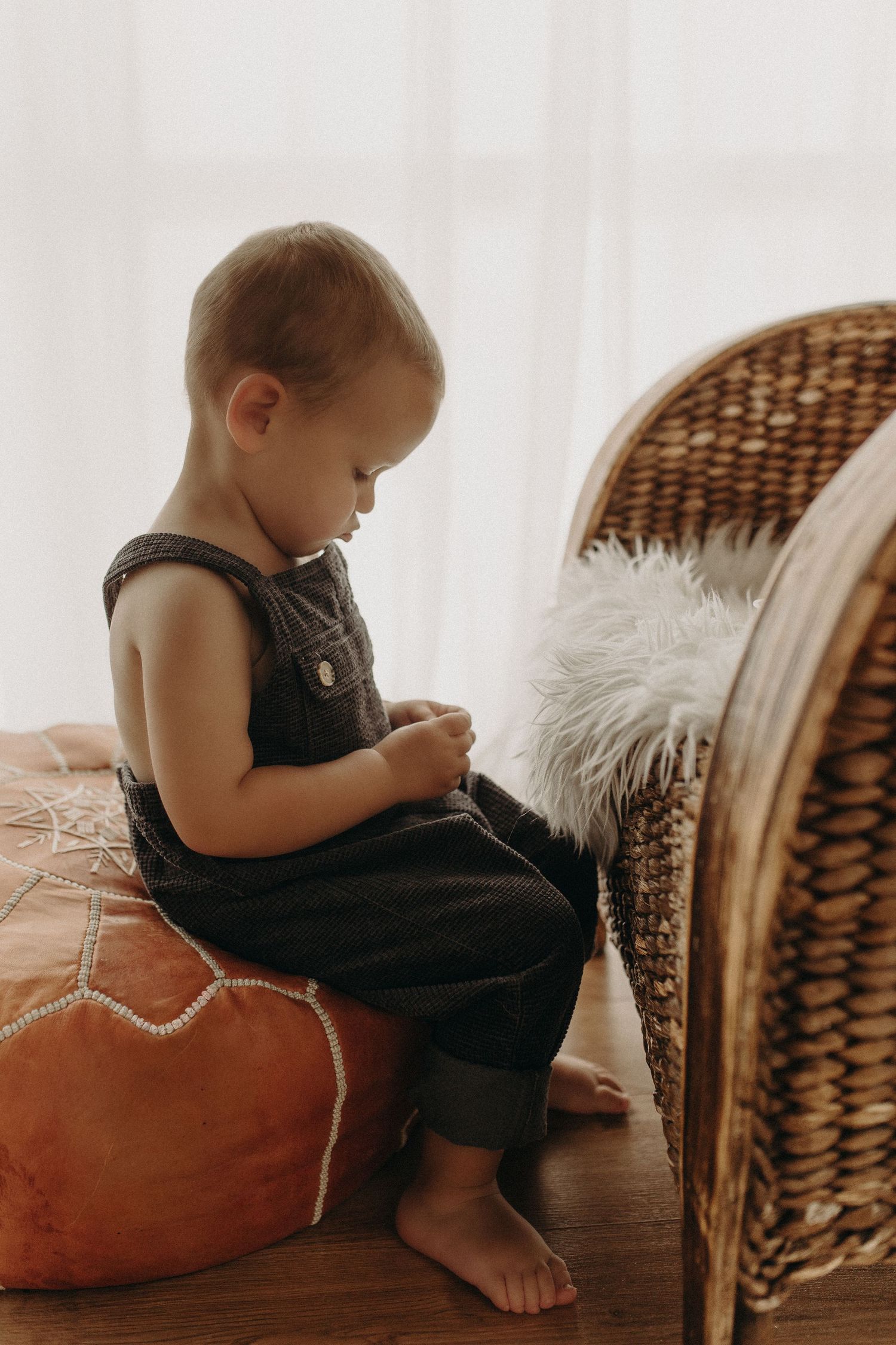 Toddler in dark overalls sitting on an orange pouf beside a wicker chair in a bright, cozy room.