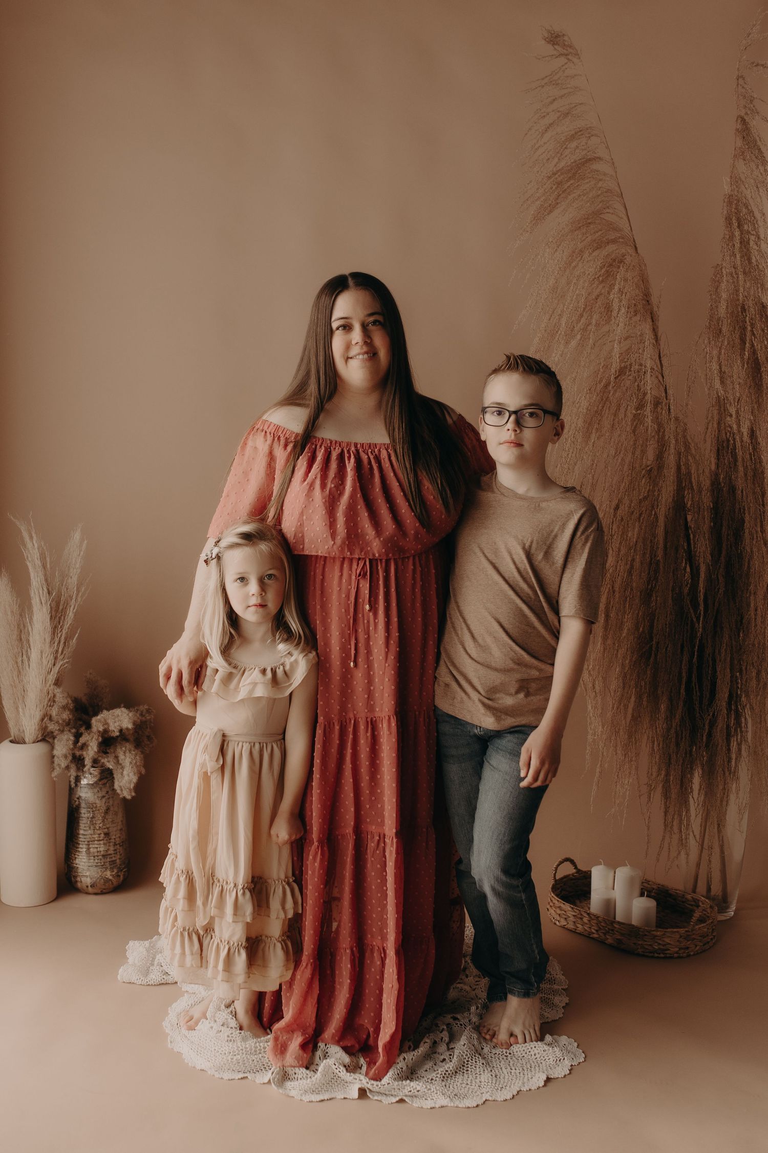 A mother in a rust off-shoulder maxi dress poses with her young daughter and son in a warm boho studio setting.