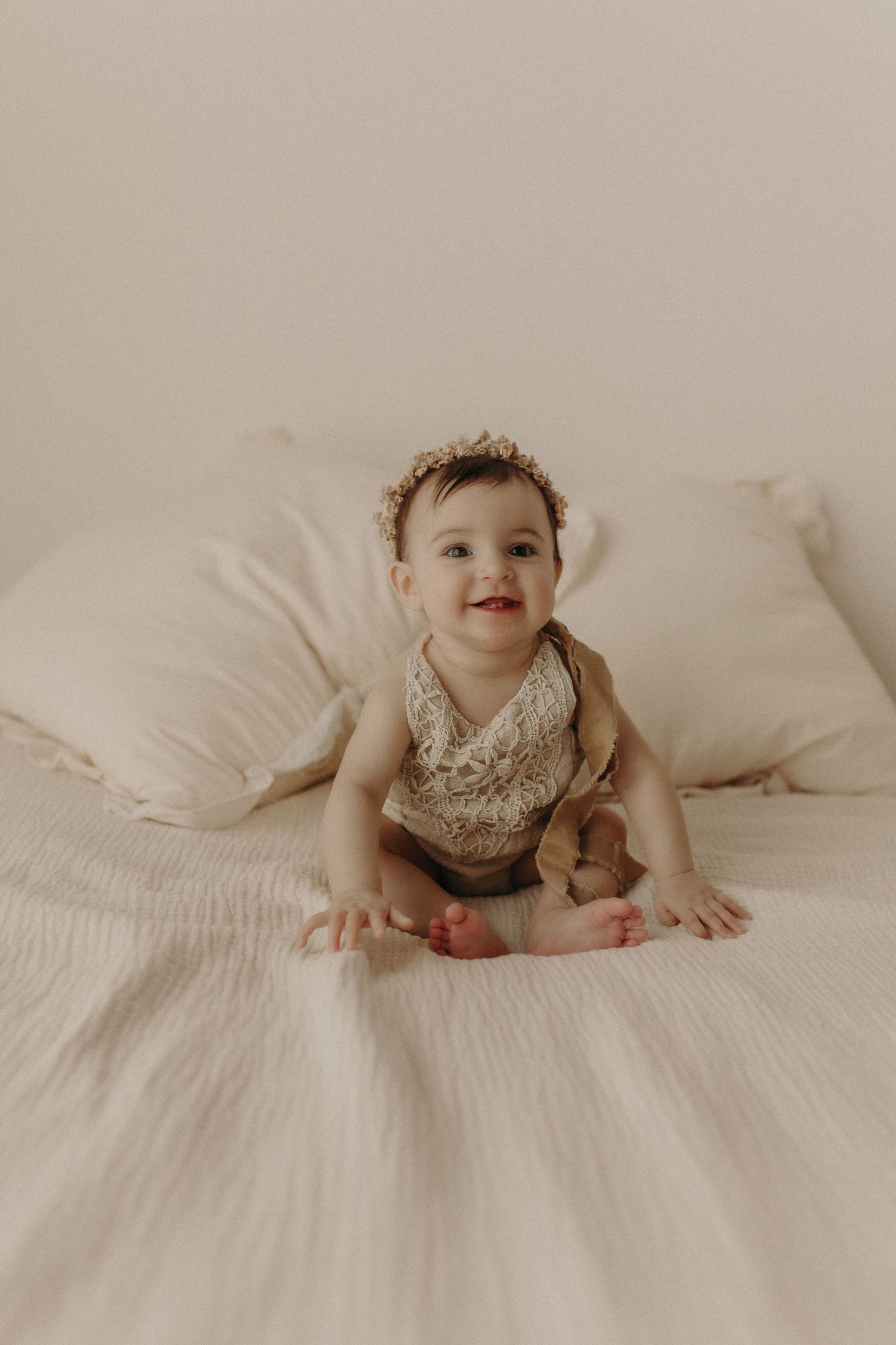 Smiling baby sitting upright on a white bed with pillows, wearing a floral bib and brown shorts.