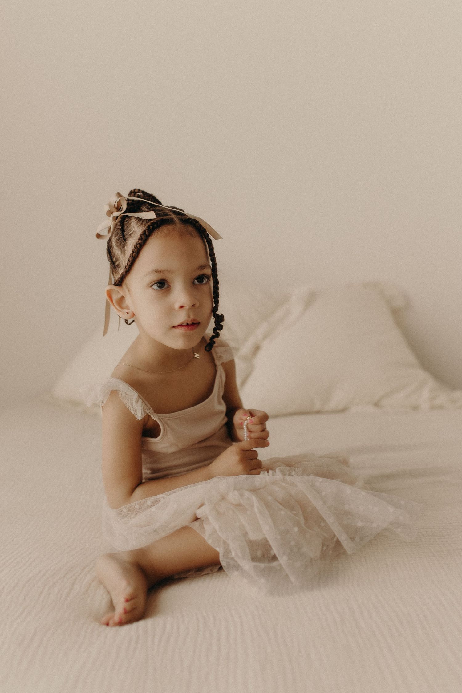 Young girl in white tulle dress with braided hair sits on a bed, looking away thoughtfully.