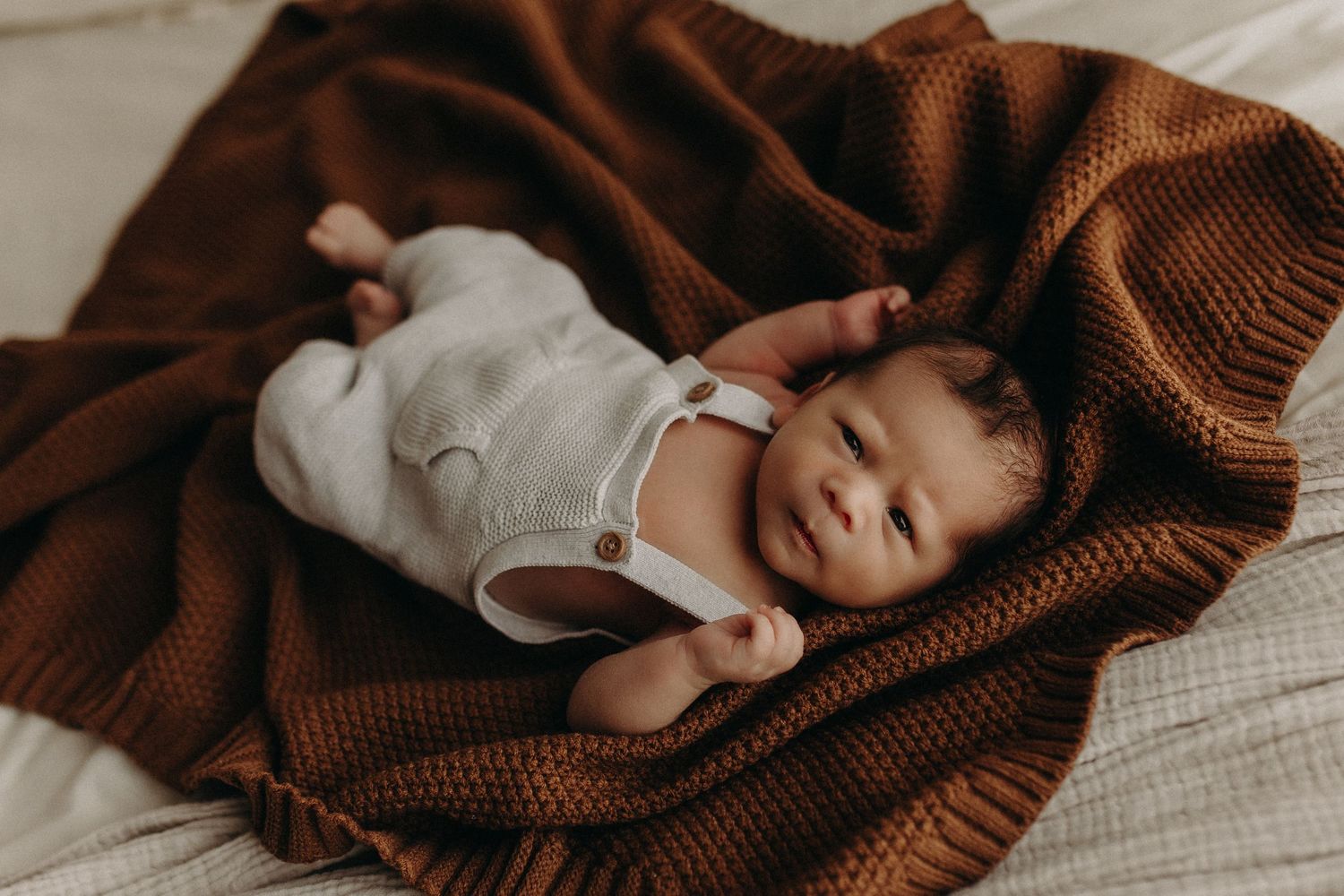 A smiling newborn baby lies on a rust-colored knit blanket wearing a white linen romper.