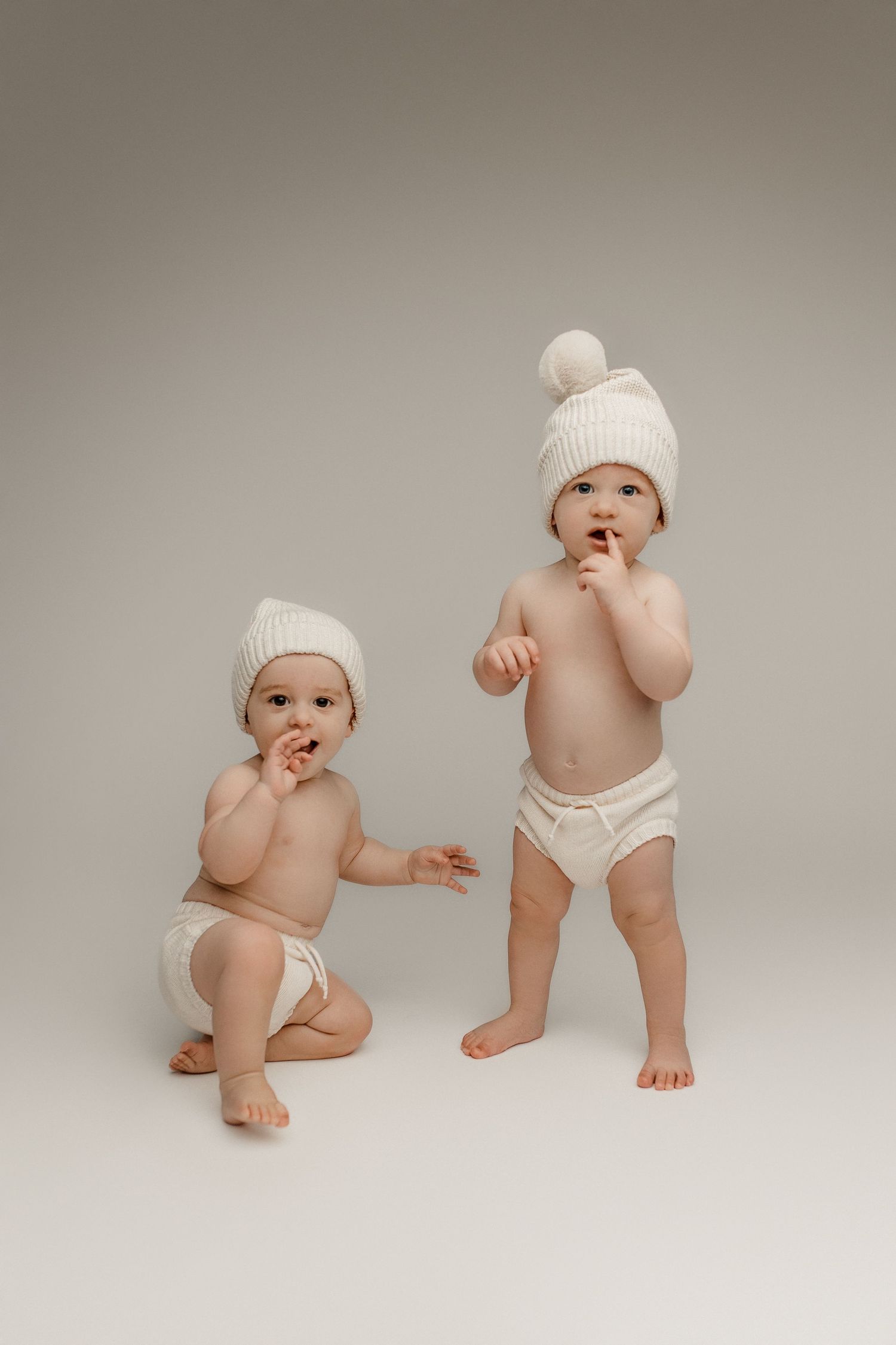 Two babies in white knit hats and diapers, one sitting and one standing, on a light gray background.