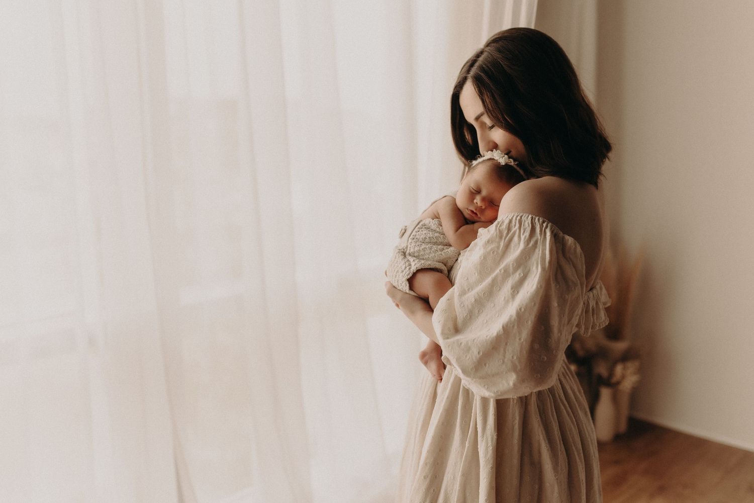 A mother in a beige off-shoulder dress tenderly holds her sleeping newborn baby near a sunlit window.