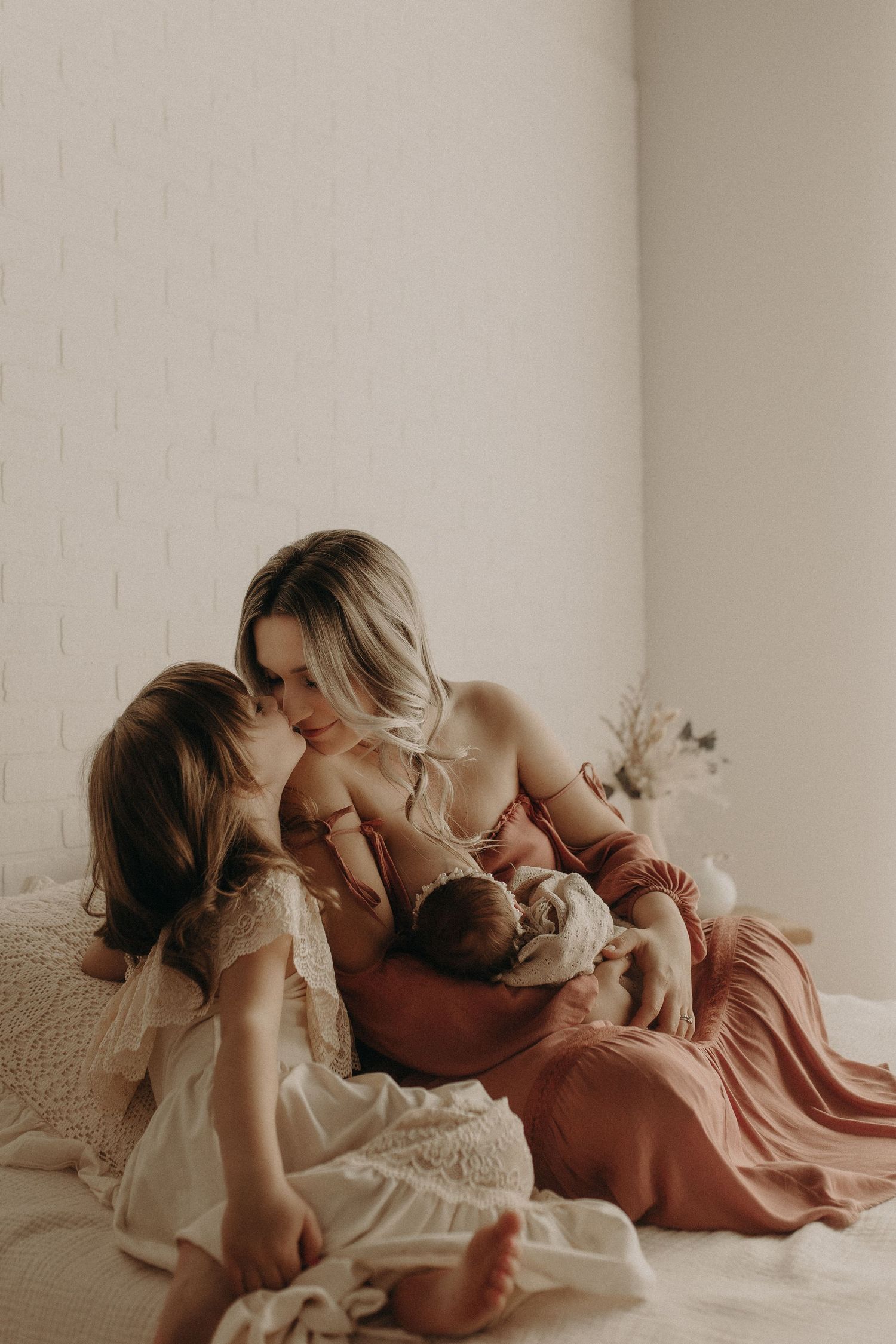 A mother in a rust dress sits on a bed breastfeeding her newborn while her young daughter leans in closely.