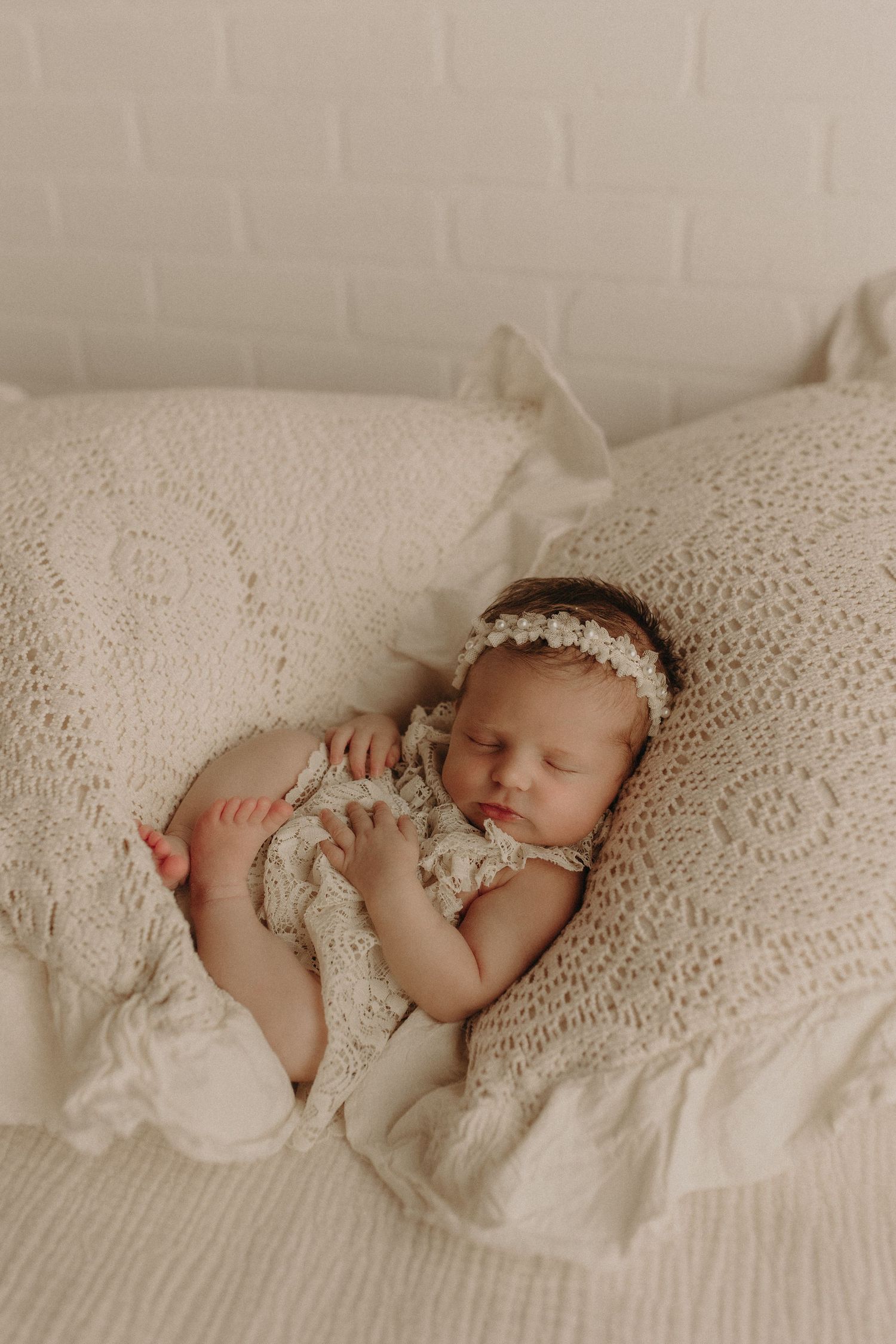 Sleeping newborn baby girl in cream lace romper and floral headband resting on white crochet pillows.
