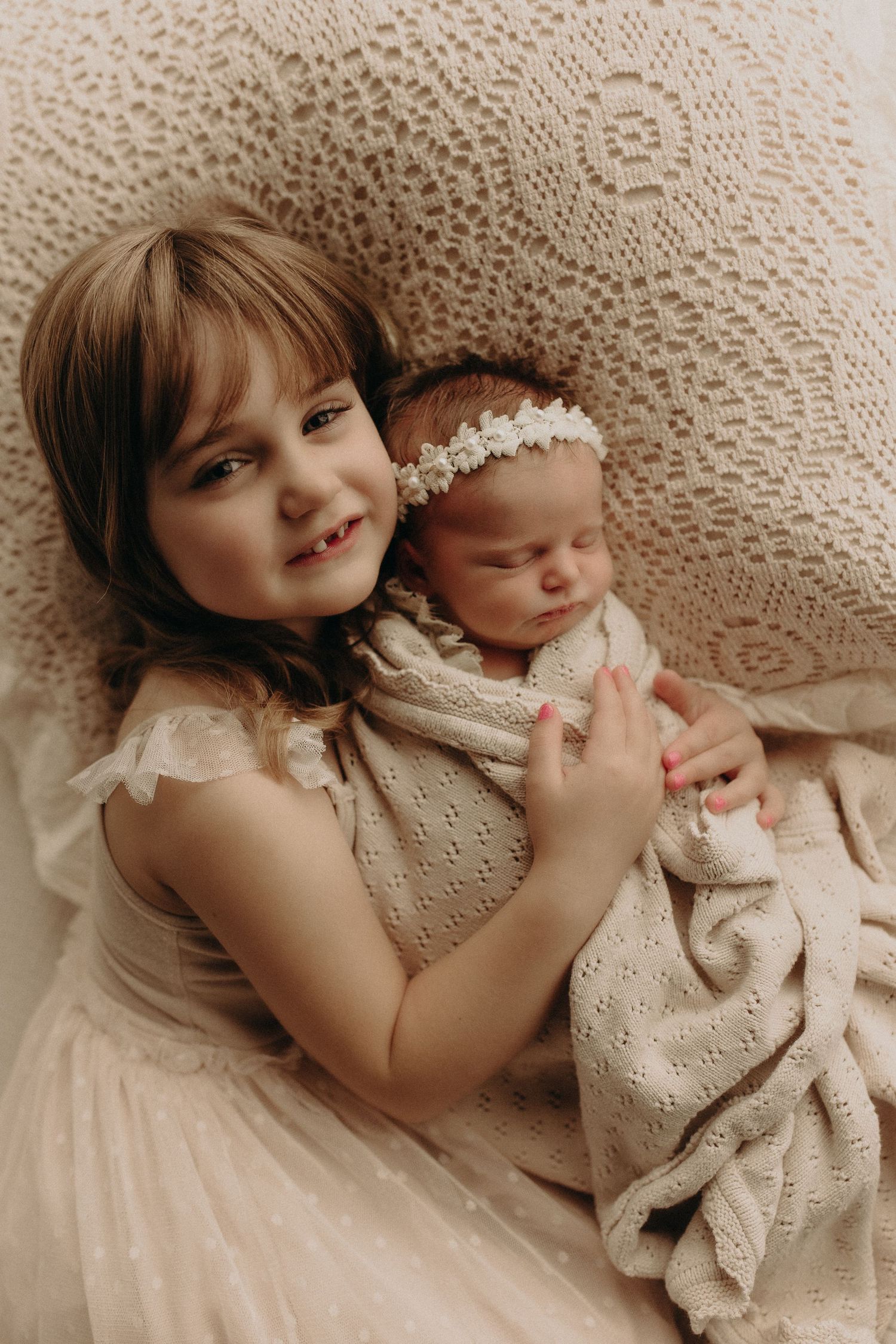 Young girl in white dress tenderly holds sleeping newborn sibling wrapped in cream blanket with floral headband.