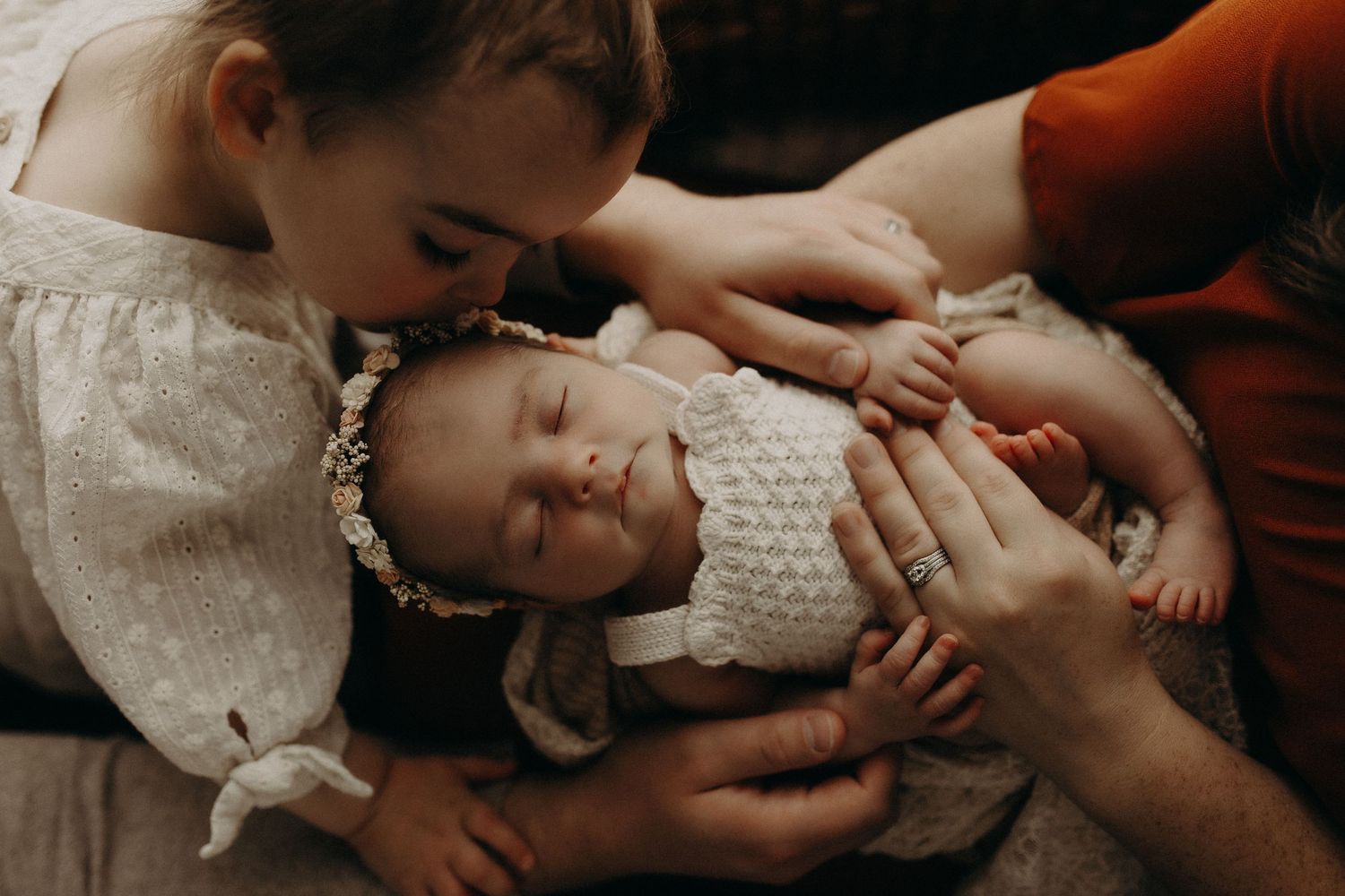A mother and young girl lovingly kiss a sleeping newborn baby dressed in a white knit outfit and floral headband.