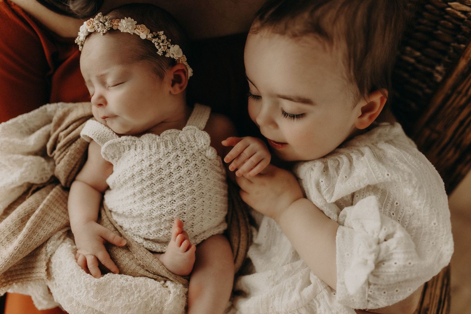 A sleeping newborn baby in a knit romper with a floral headband cuddles beside an older toddler sibling.