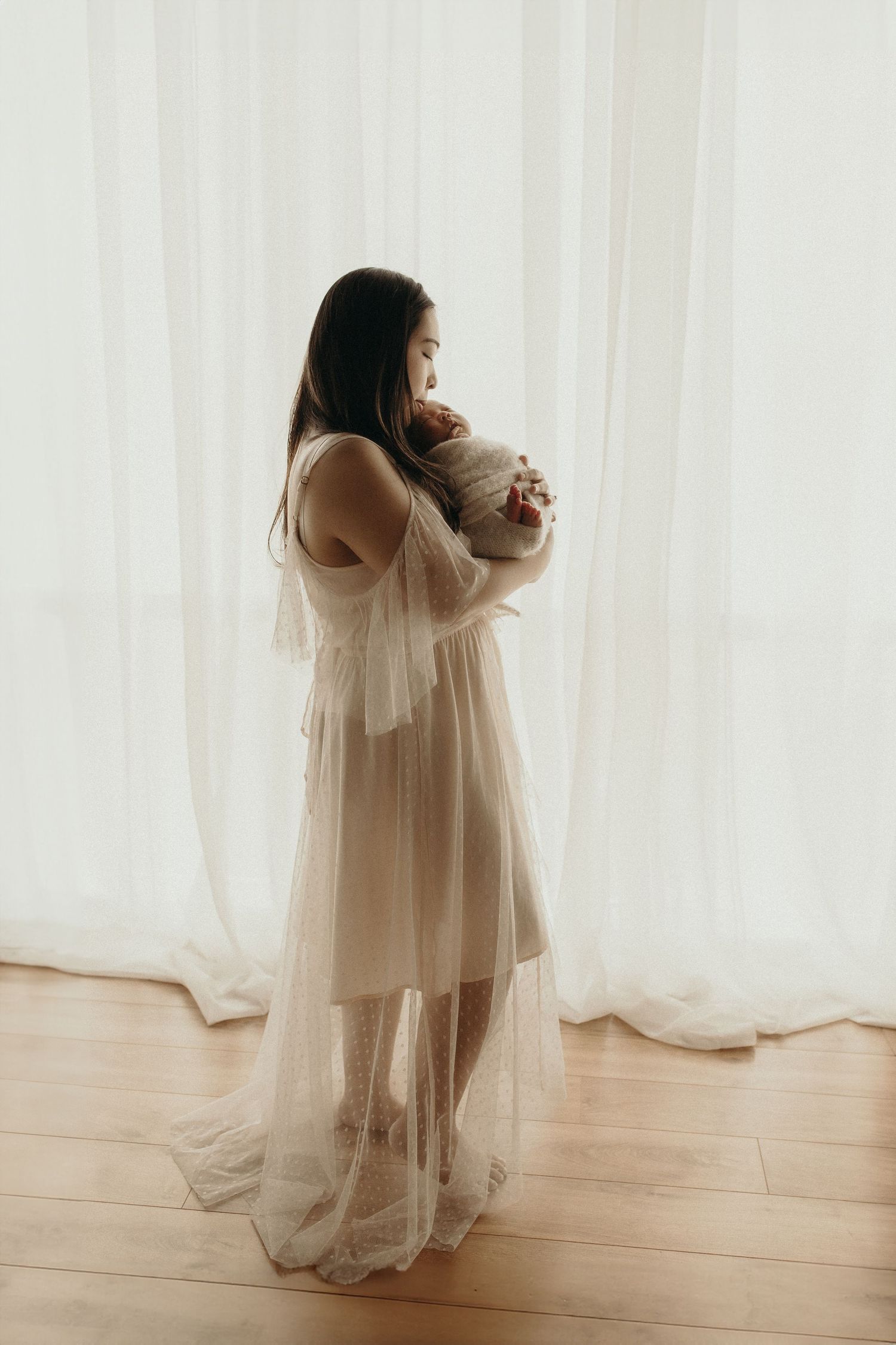 A mother in a sheer white lace gown cradles her newborn baby by a sunlit window on a hardwood floor.