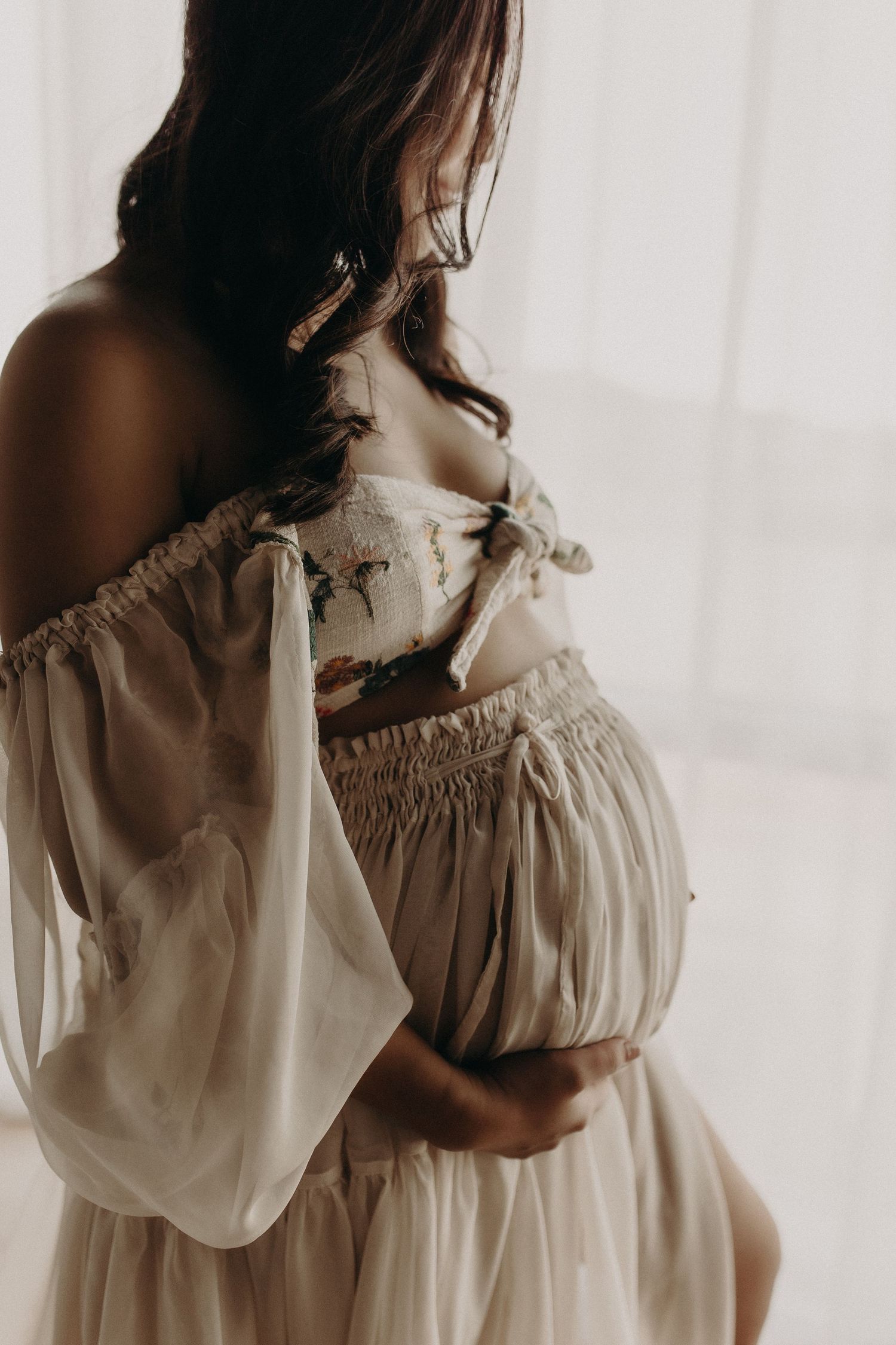 Pregnant woman in elegant off-shoulder cream floral dress cradling baby bump near window.