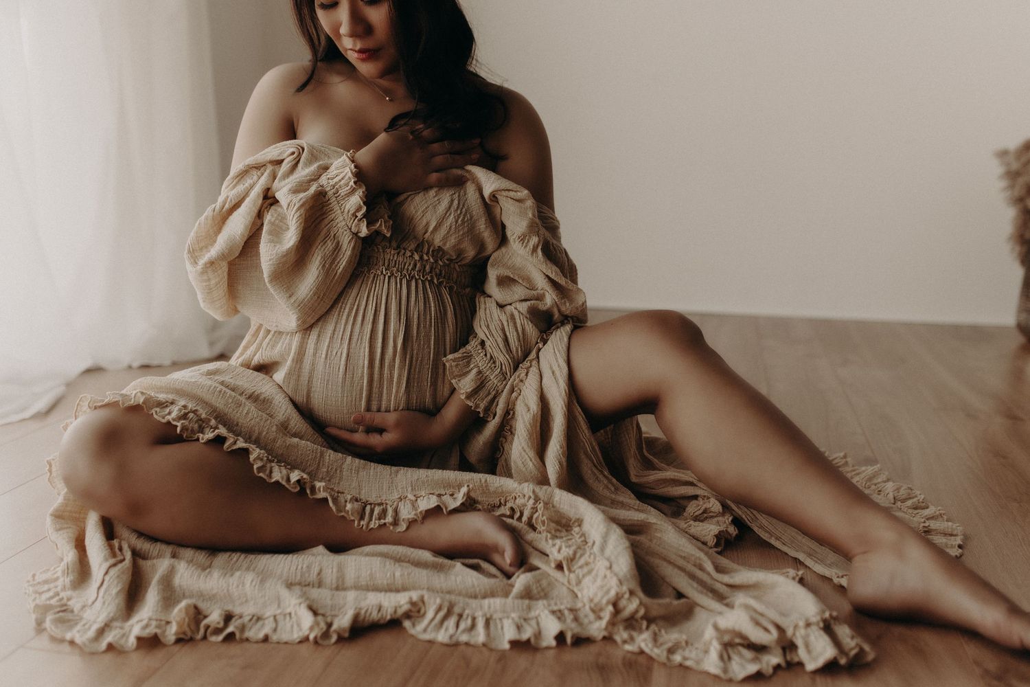 Pregnant woman in a flowing beige dress sitting gracefully on a rug, cradling her baby bump in a soft, natural light setting.