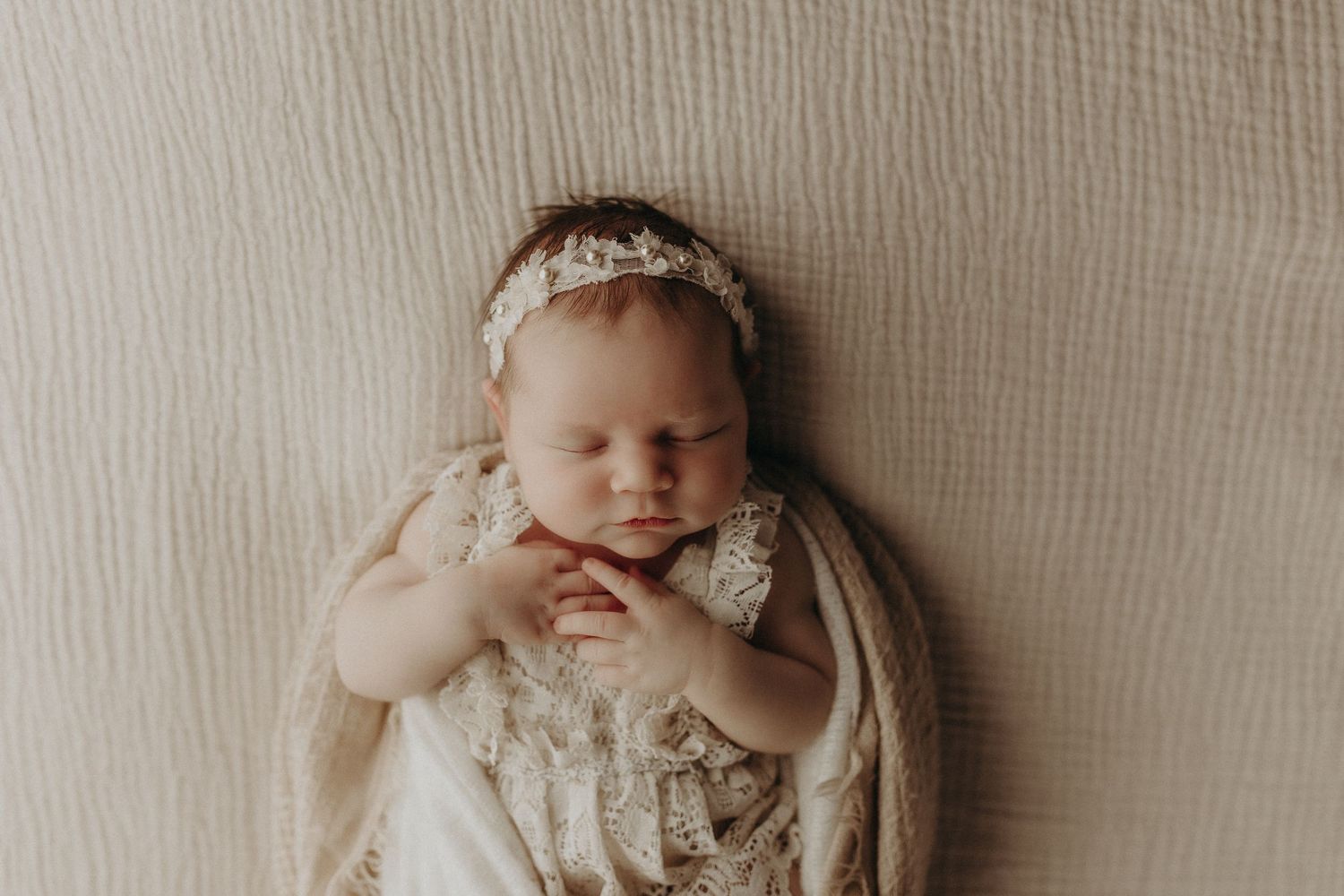 Peaceful newborn baby sleeping wrapped in cream lace swaddle with floral headband on soft white background.