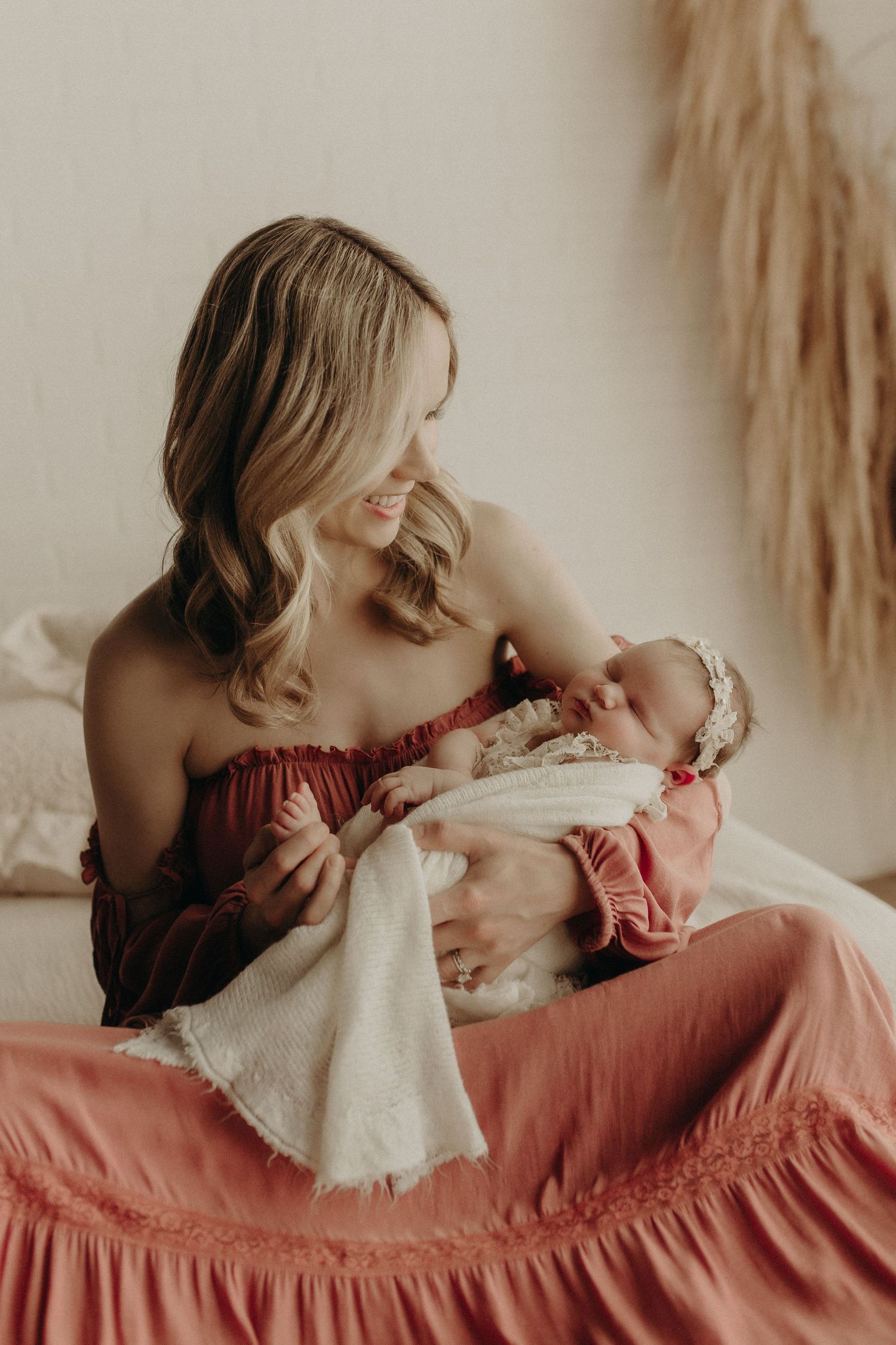 A young mother in a rust-red dress tenderly holds her newborn baby, smiling softly in a warm, neutral-toned room.
