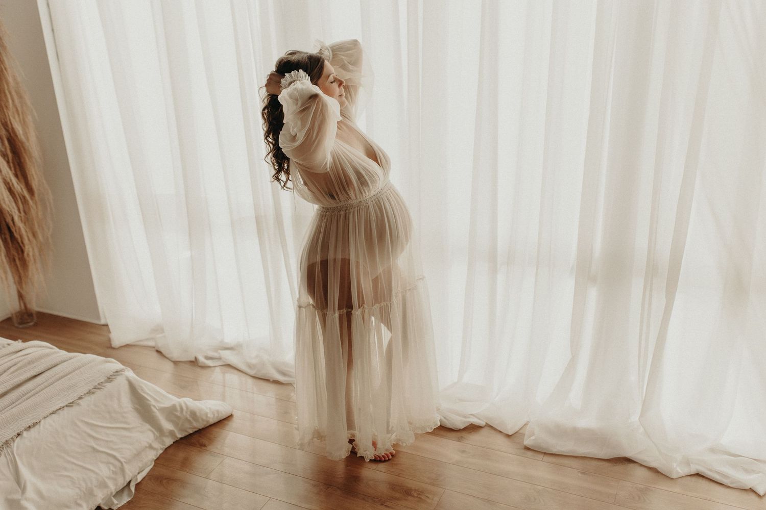 Pregnant woman in sheer white maternity gown standing by curtained window in soft natural light.
