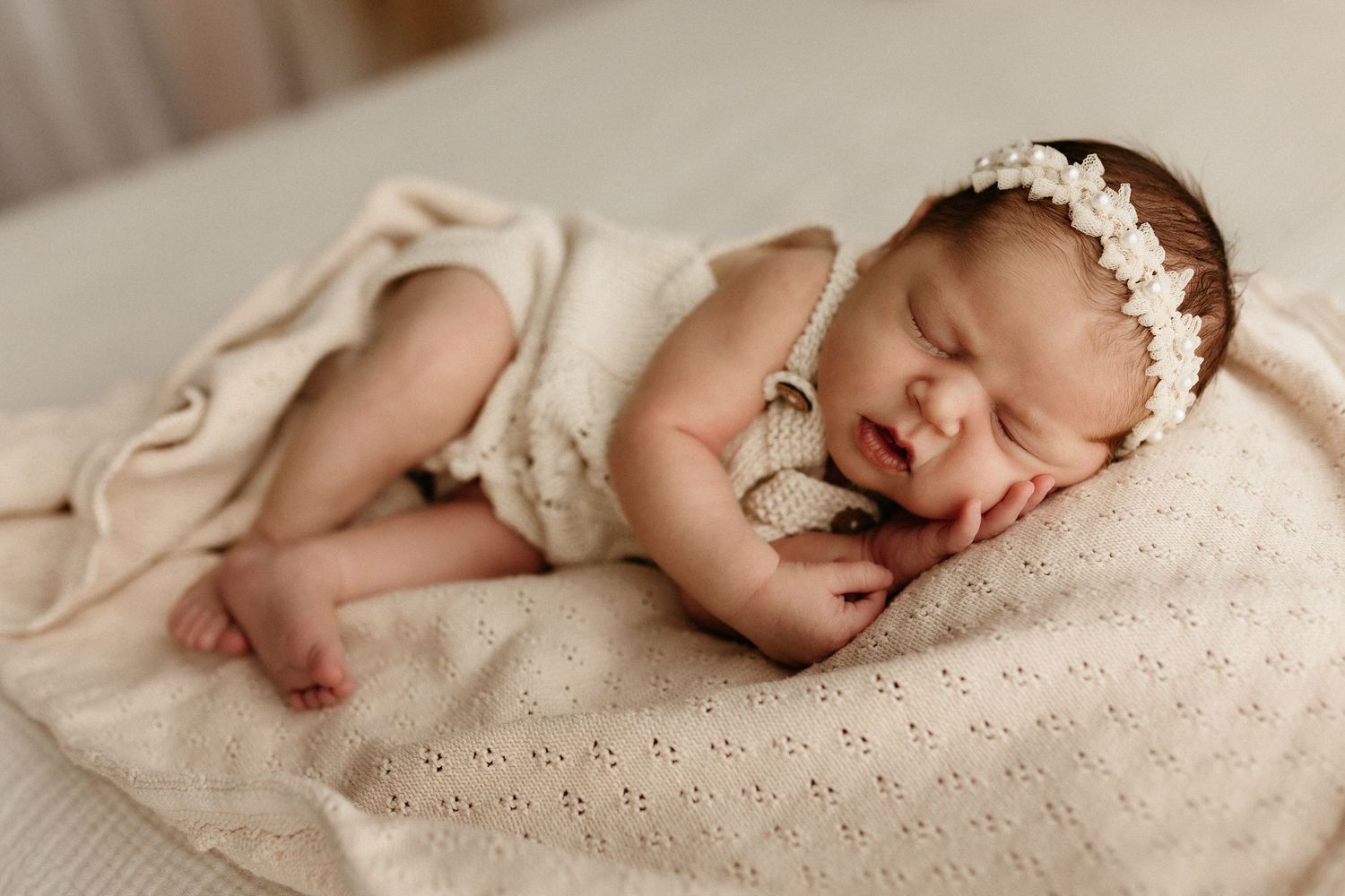 Peaceful newborn baby girl sleeping on a cream blanket wearing a delicate floral headband in a soft studio setting.