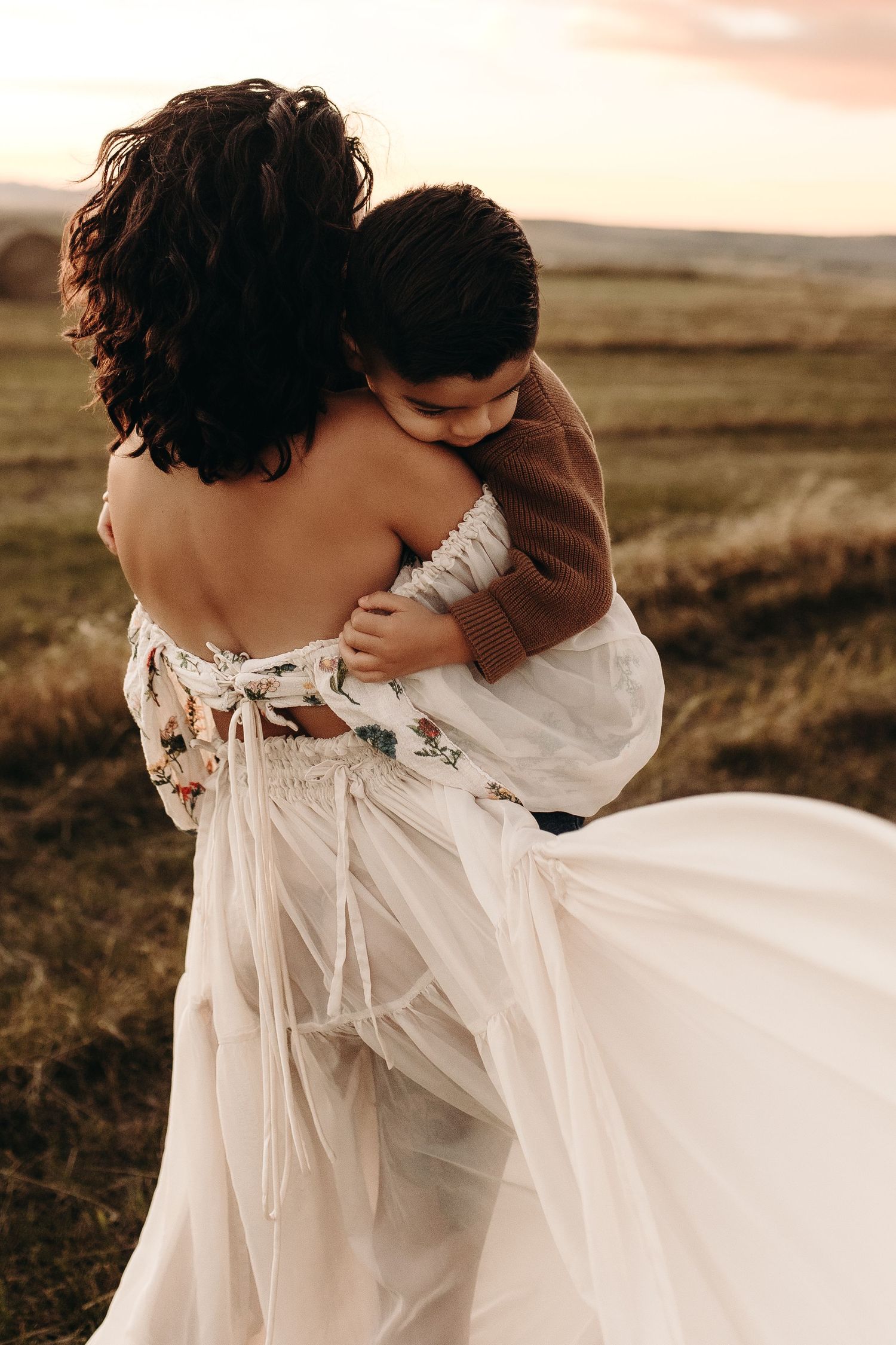 A woman in a flowy white floral dress holds a young child in a warm embrace outdoors at sunset.