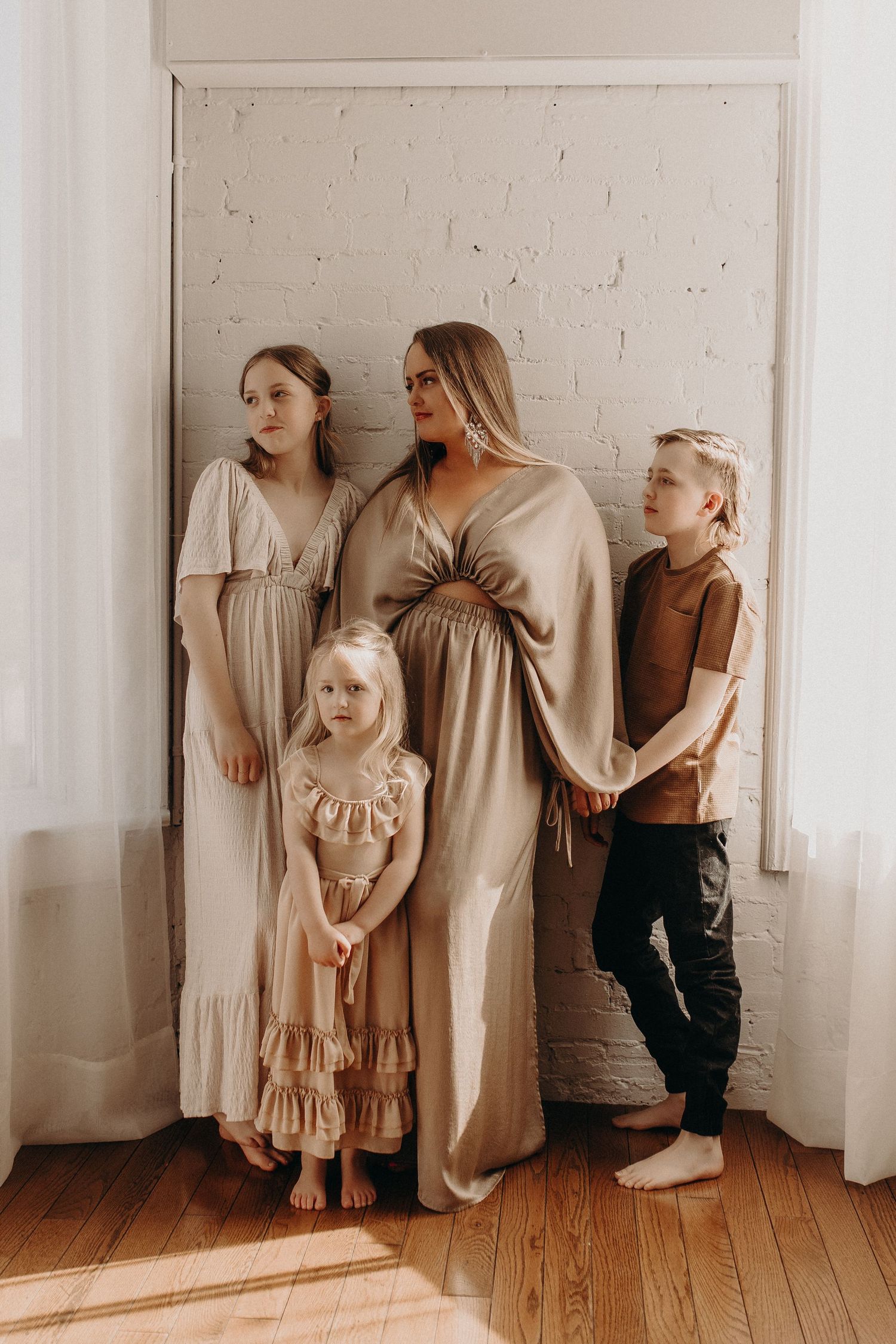 Pregnant woman in gold dress poses with three children against white brick wall in warm, natural light.
